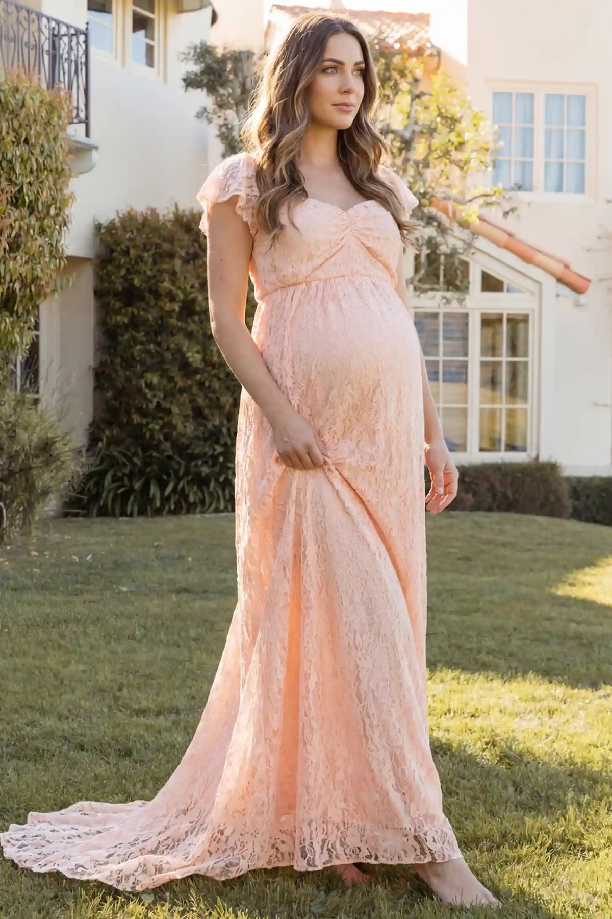 Lace Off Shoulder Maternity Maxi Dress in blush pink featuring a sweetheart neckline with a ruched bust and a full-length skirt, shown on a pregnant woman outdoors on a lawn, side view.