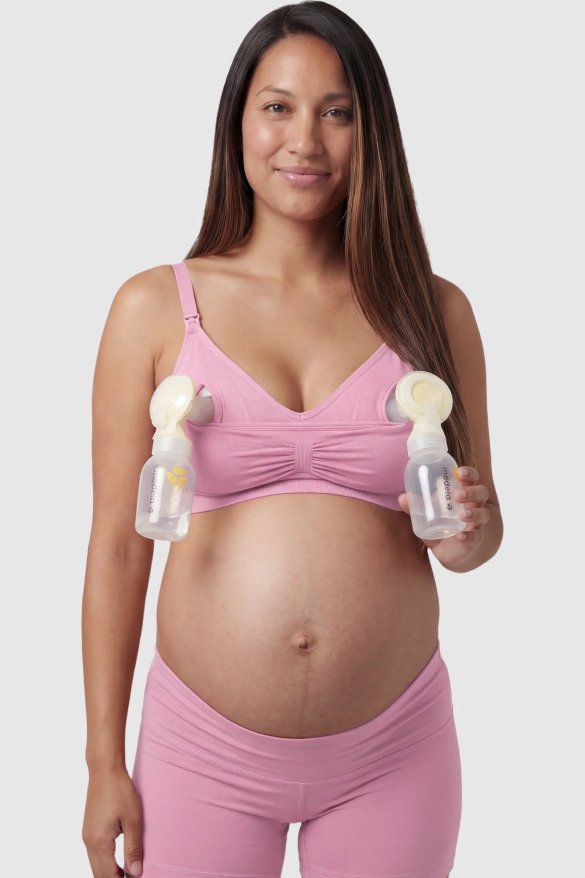 Seamless Ruched Front Maternity Nursing Bra in peach pink with hands-free pumping bottles attached, showing the innovative crossover design on a smiling pregnant model showing front.