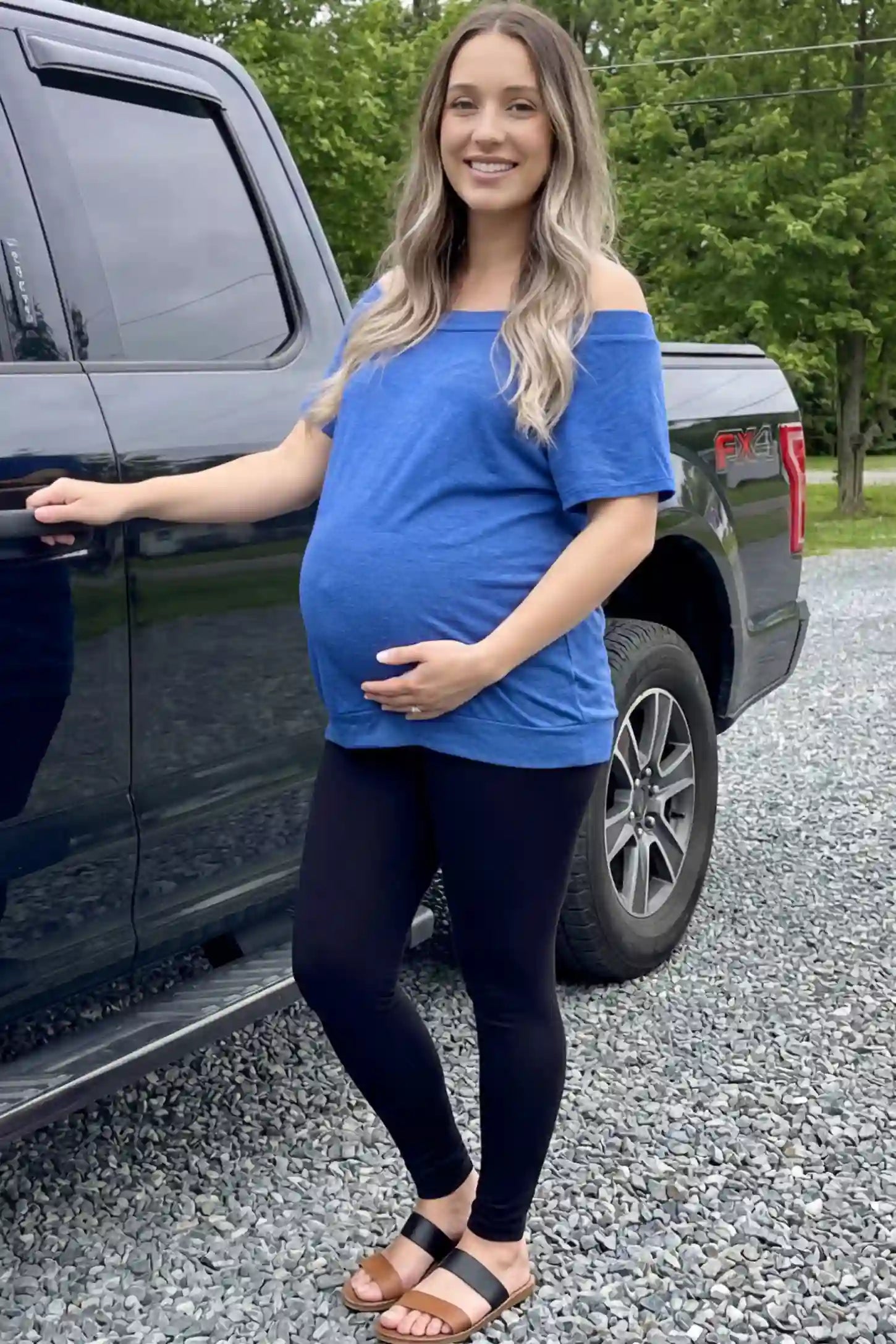 Convertible Neck Short Sleeve Maternity Top in blue featuring an adjustable neckline and a comfortable banded hem paired with black leggings worn by a pregnant woman standing next to a truck, front view.