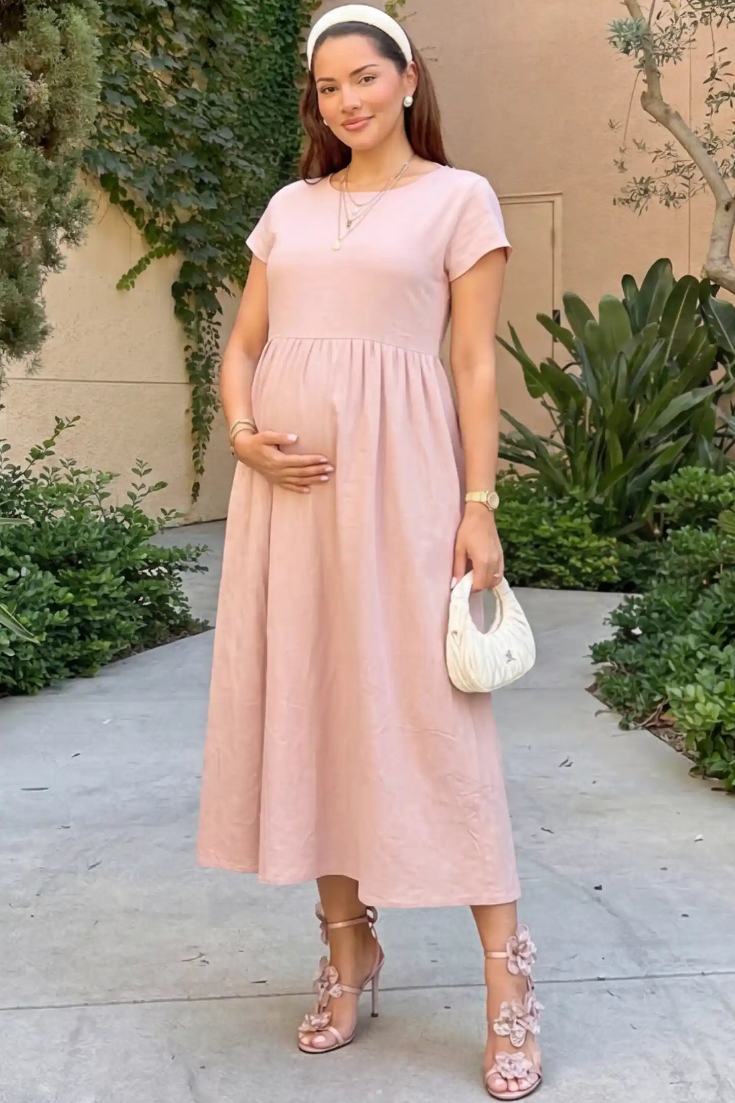 Crew Neck Short Sleeve Maternity Midi Dress in Pink featuring a flowy shift silhouette on a pregnant woman wearing a white headband, standing on an outdoor walkway, front view.