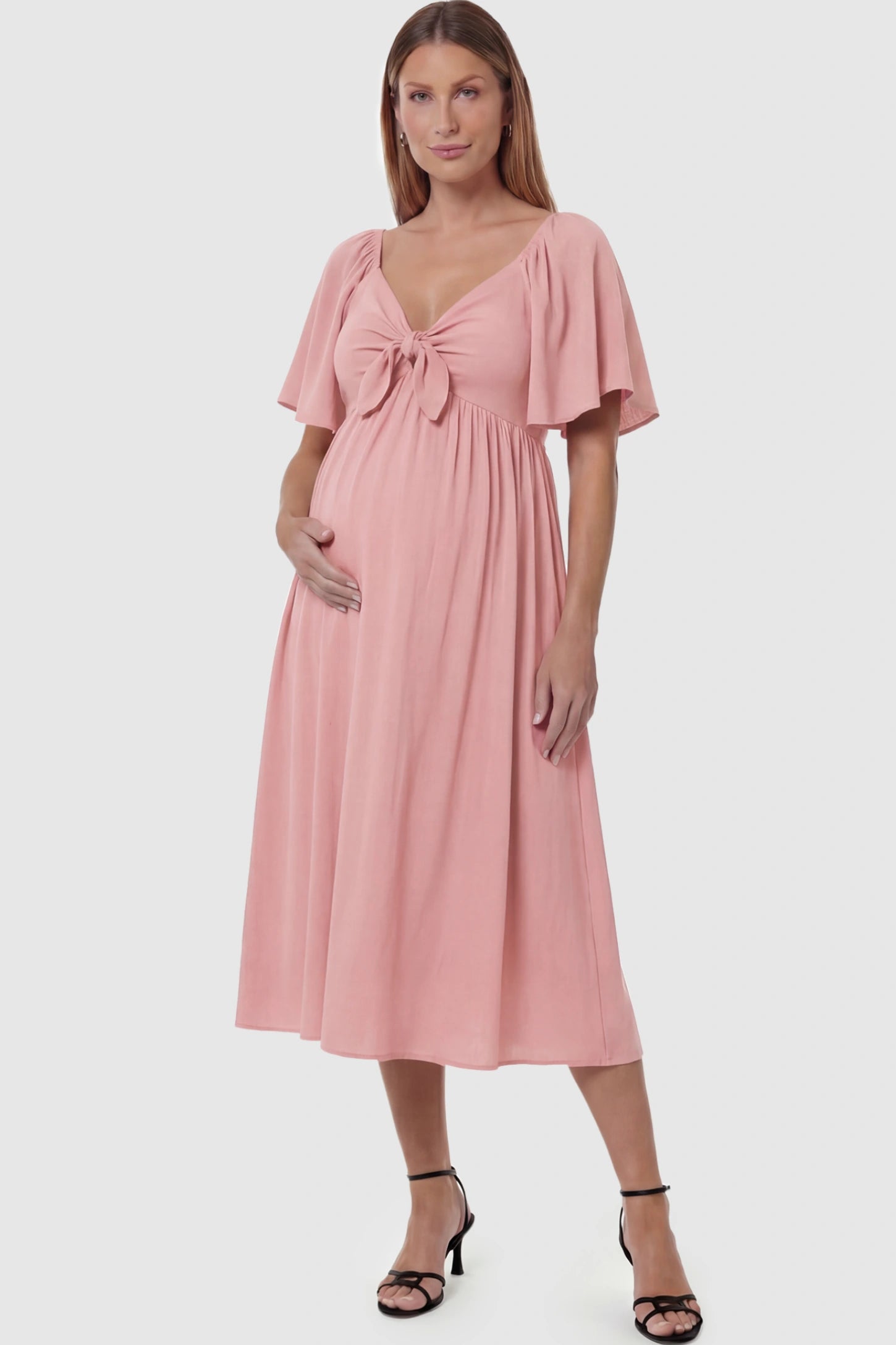 Front Tie Flutter Sleeve Maternity Midi Dress in pink featuring a knotted V-neckline and flowy skirt on a pregnant model posing front.