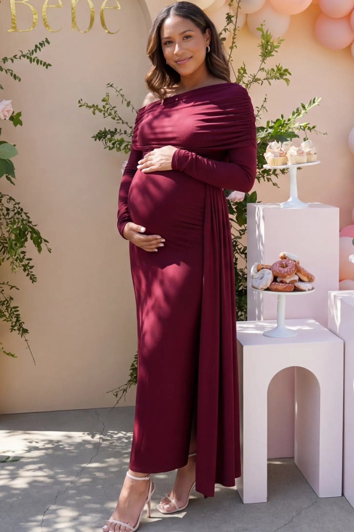 Off Shoulder Draped Wrap Bodycon Maternity Maxi Dress in Wine Red featuring side ruching and long sleeves on a pregnant model cradling her baby bump at a baby shower, front view.