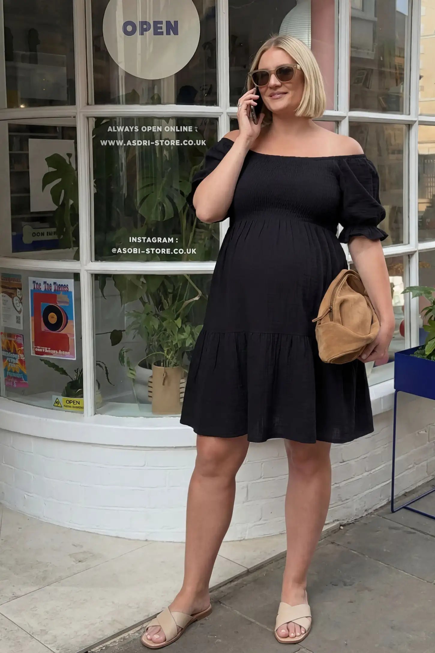 Smocked off-shoulder empire waist maternity short dress in black with smocked bodice detail, front view on pregnant model