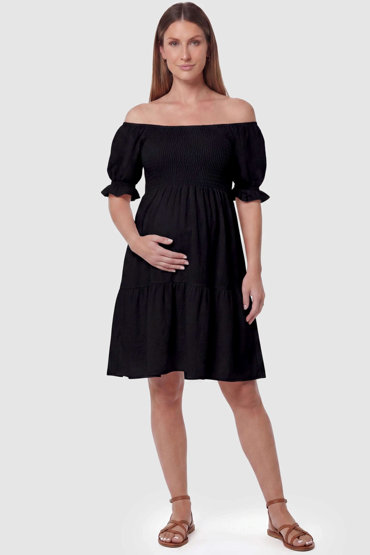 Smocked off-shoulder empire waist maternity short dress in black with smocked bodice detail, front view on pregnant model