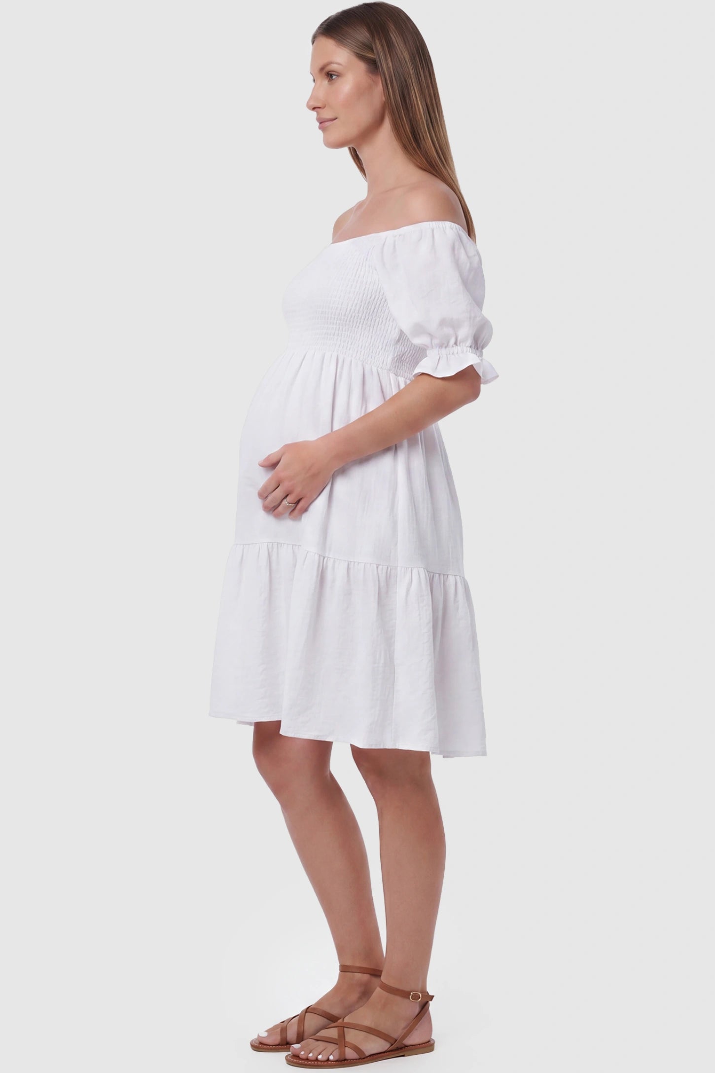 Smocked Off-Shoulder Empire Waist Maternity Short Dress Smocked off-shoulder empire waist maternity short dress in white with bump-friendly empire waist fit, side view on pregnant model