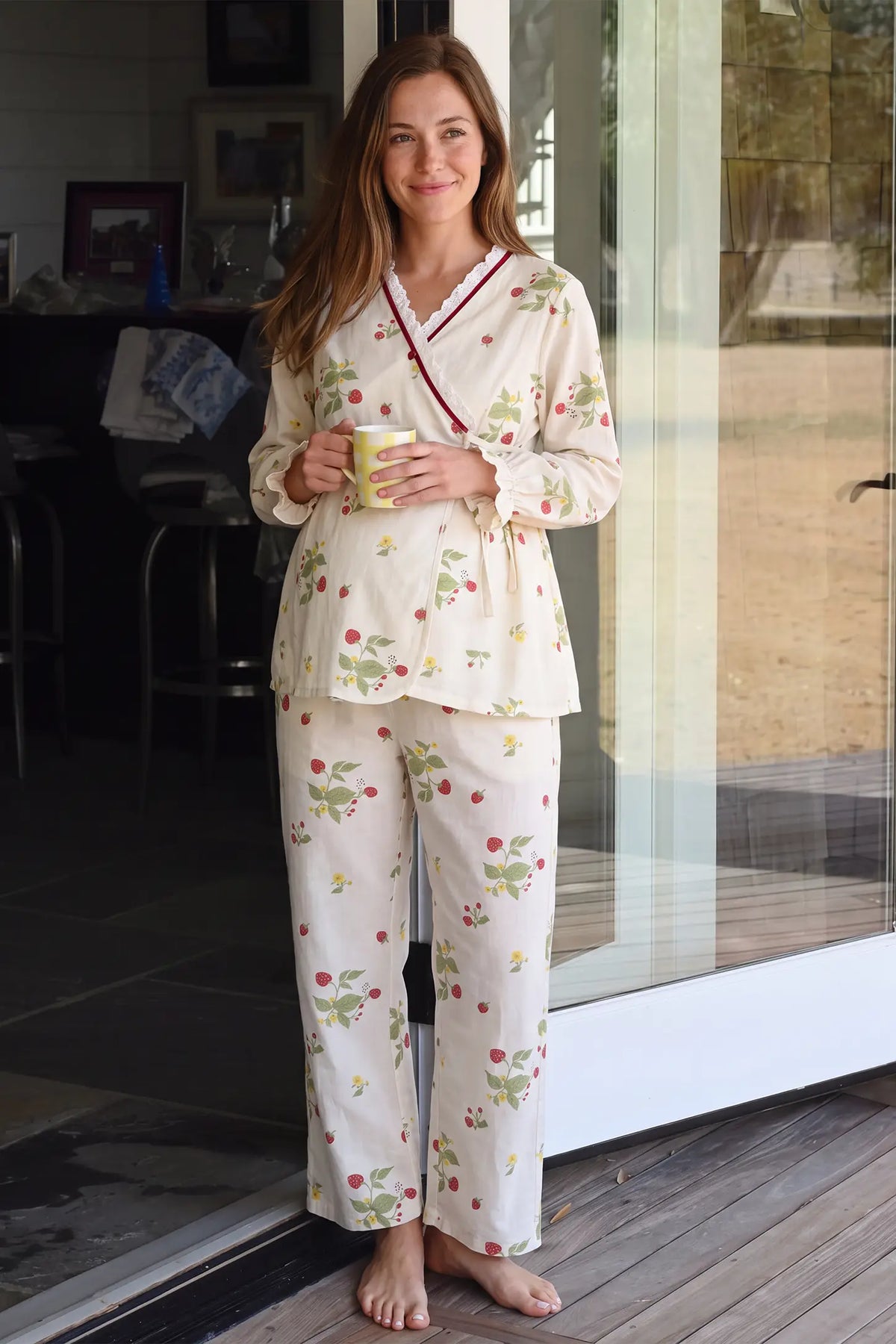 Cotton Long Sleeve Top & Pants Maternity Pajama Set Full-length lifestyle shot of a smiling pregnant model standing near a sunlit doorway wearing the strawberry pattern cotton long sleeve maternity pajama set while holding a cup.