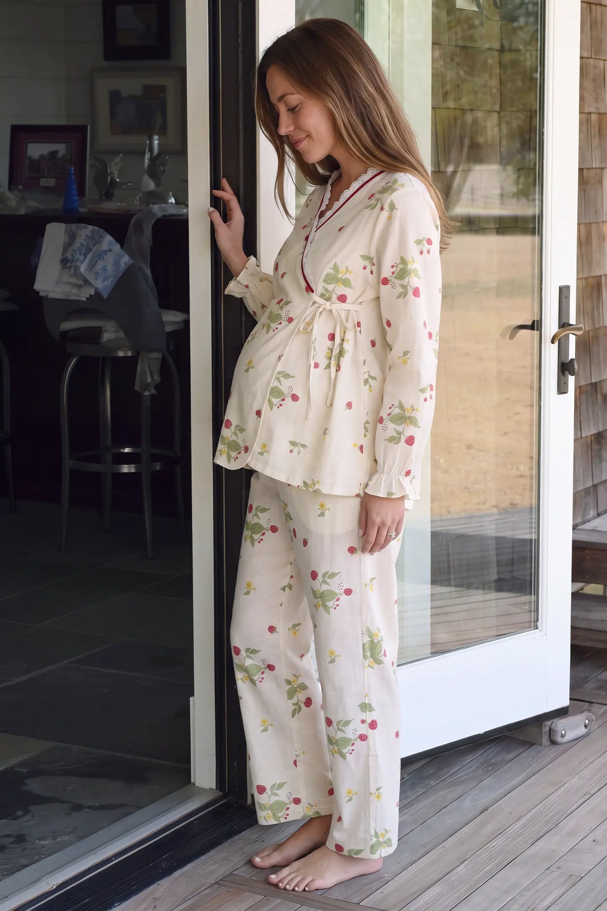 Cotton Long Sleeve Top & Pants Maternity Pajama Set Three-quarter side view lifestyle photo of a pregnant model standing by an open glass door in the cozy strawberry long-sleeve maternity pj set.