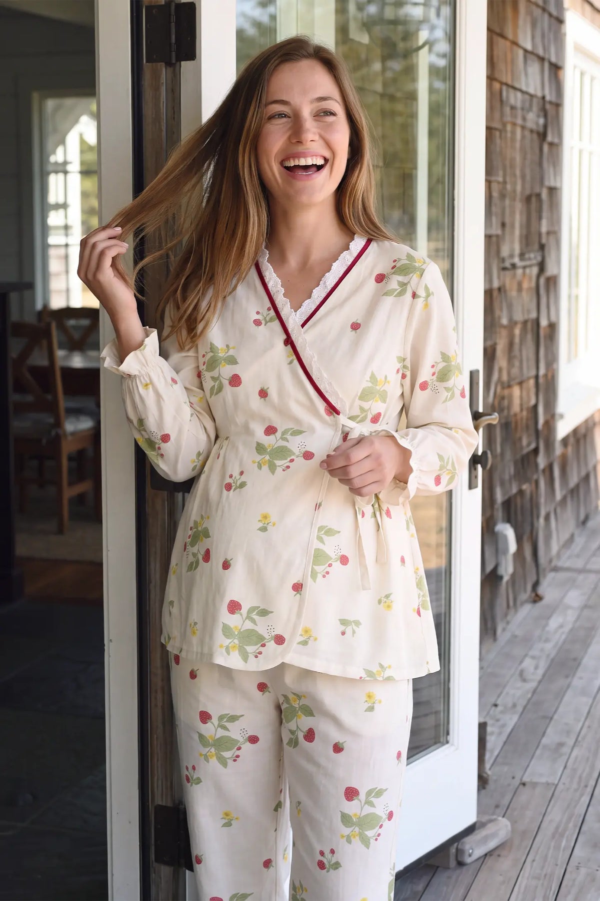 Cotton Long Sleeve Top & Pants Maternity Pajama Set Medium shot of a smiling pregnant model indoors near natural light showing the v-neckline and side-tie wrap detail of the strawberry print maternity pajamas.