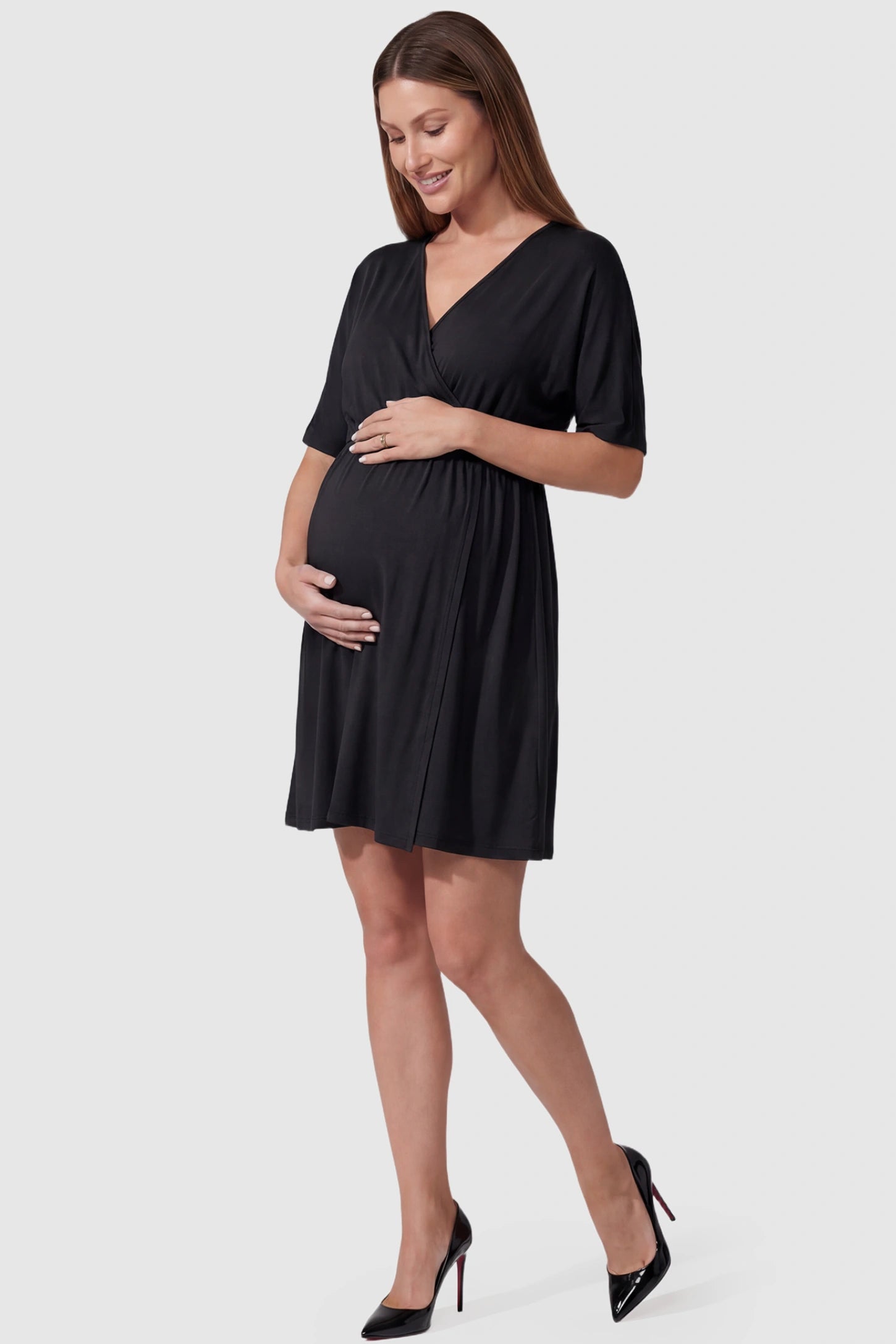 Wrap Front Empire Waist Maternity & Nursing Mini Dress Wrap Front Empire Waist Maternity & Nursing Dress in black featuring a draped crossover V-neck and short sleeves on pregnant model front view.