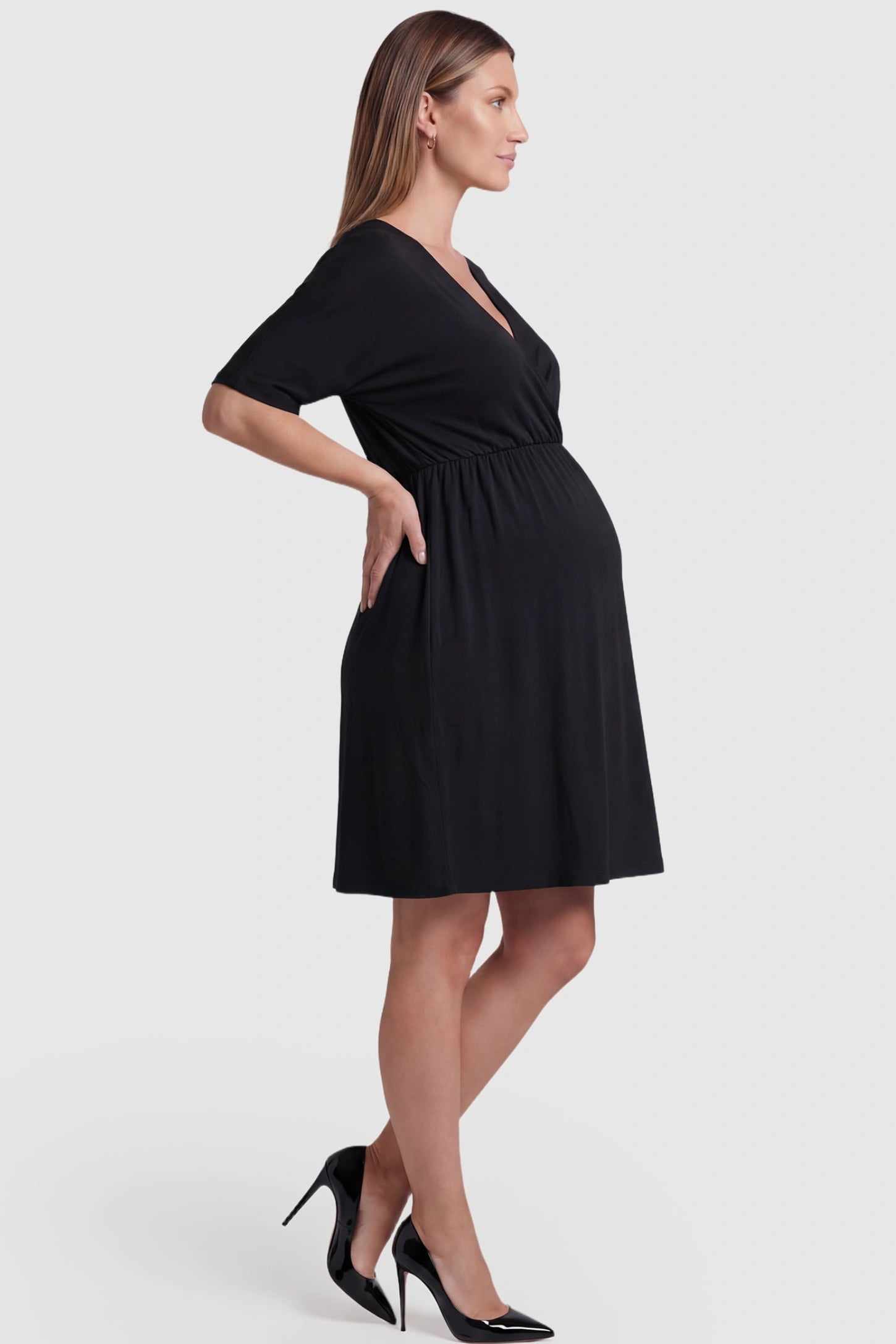 Wrap Front Empire Waist Maternity & Nursing Mini Dress Wrap Front Empire Waist Maternity & Nursing Dress in black featuring a draped crossover V-neck and short sleeves on pregnant model side view.