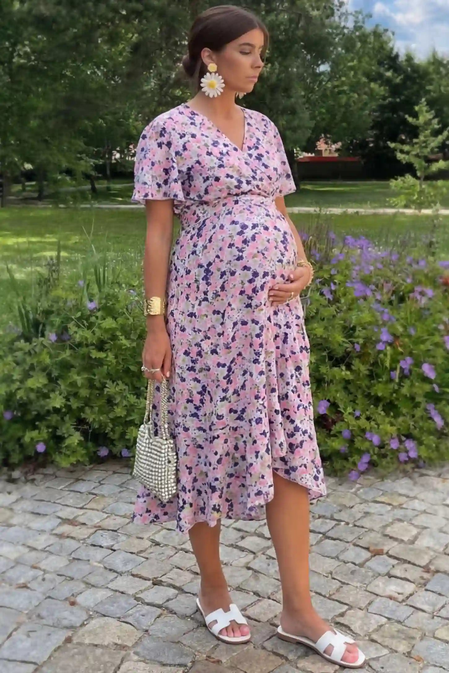 Floral V-neck ruffled hem midi maternity dress in apricot featuring V-neckline and tie waist in full body front view on a pregnant woman in outdoor setting, side view.