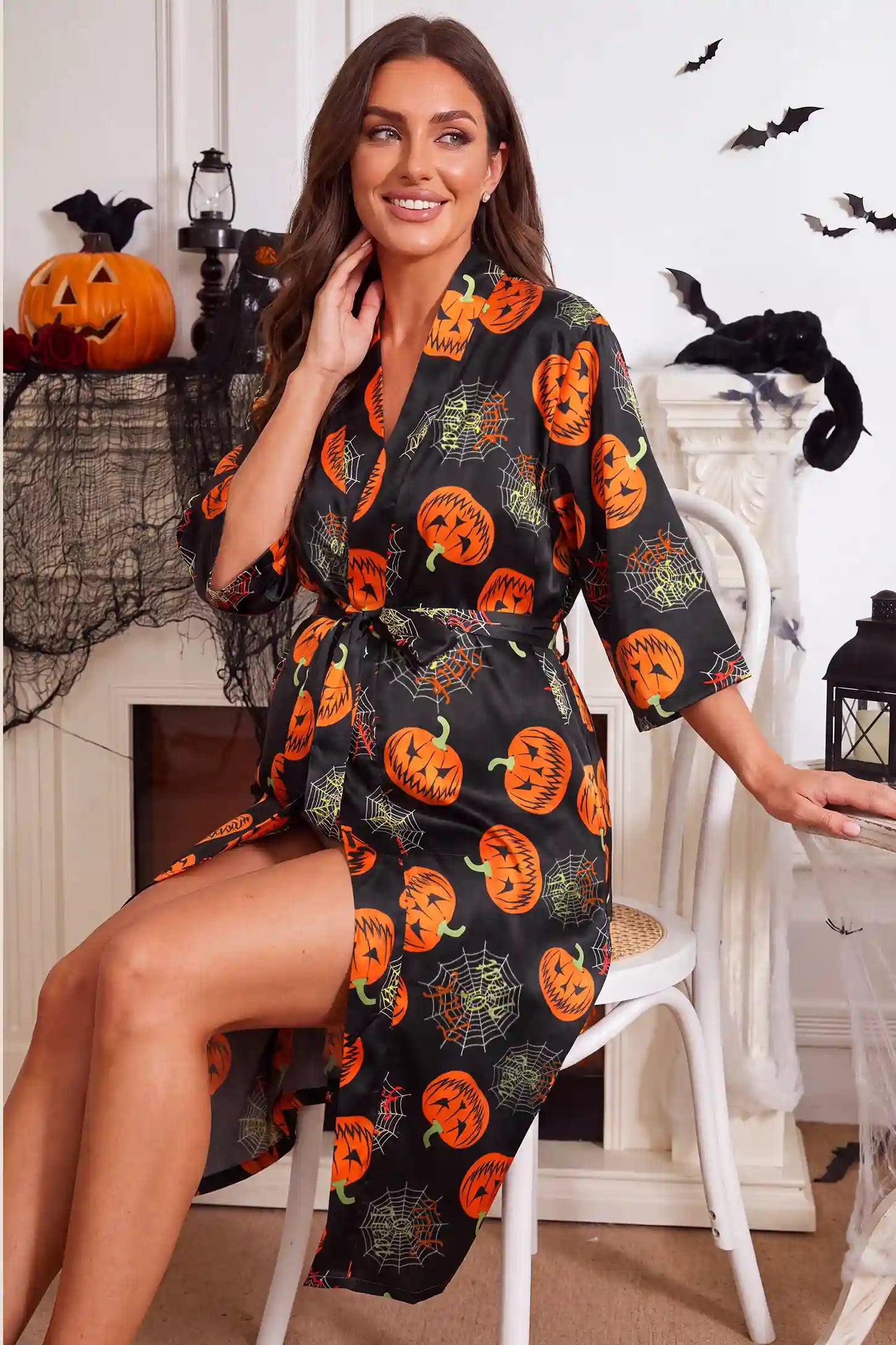 Halloween Pumpkin Print Satin Maternity Lounge Robe Halloween Pumpkin Print Satin Maternity Lounge Robe in Midnight Pumpkin featuring festive pumpkin pattern print, soft stretch fabric, waist tie sash, and relaxed fit on a pregnant model, seated front view.