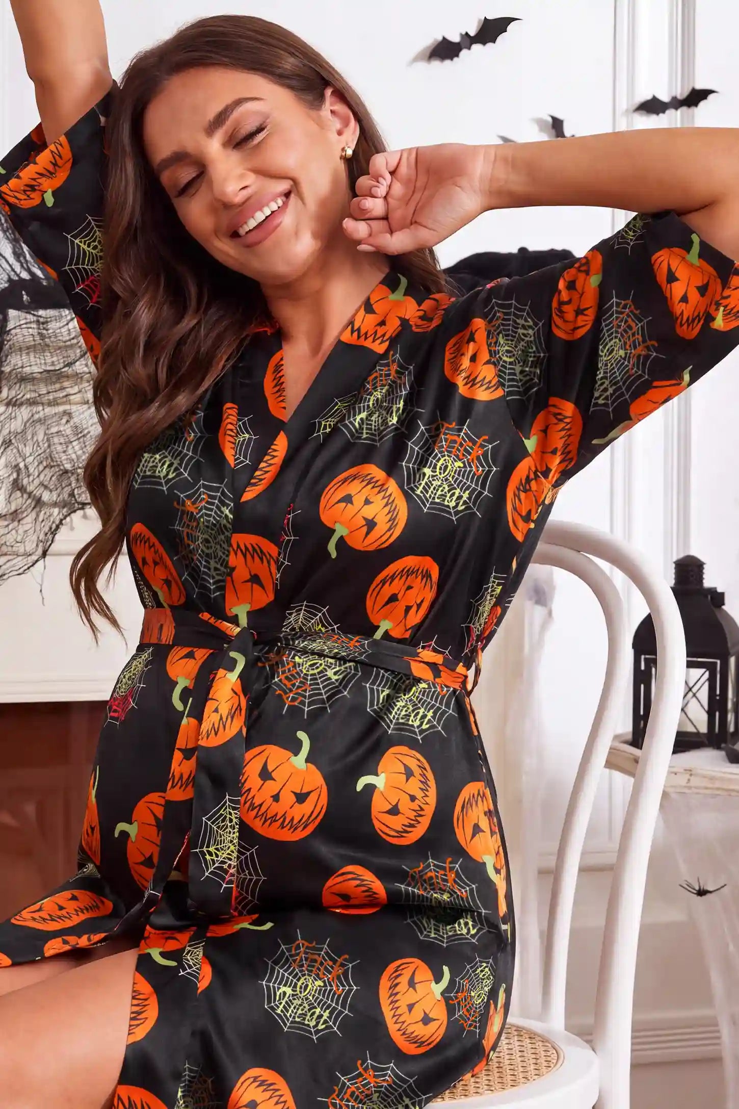 Halloween Pumpkin Print Satin Maternity Lounge Robe Halloween Pumpkin Print Satin Maternity Lounge Robe in Midnight Pumpkin featuring festive jack-o'-lantern pattern, gentle bump coverage, and easy wrap design on a pregnant model, seated close-up view.