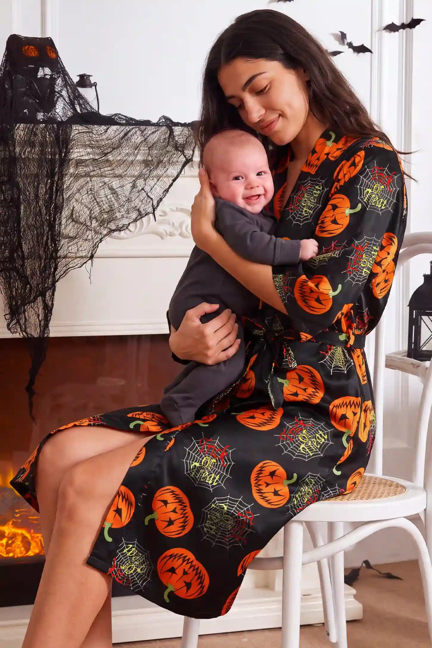 Halloween Pumpkin Print Satin Maternity Lounge Robe Halloween Pumpkin Print Satin Maternity Lounge Robe in Midnight Pumpkin featuring soft stretch material, easy wrap style, versatile postpartum design, and cozy fit on a mother holding her baby, seated view.