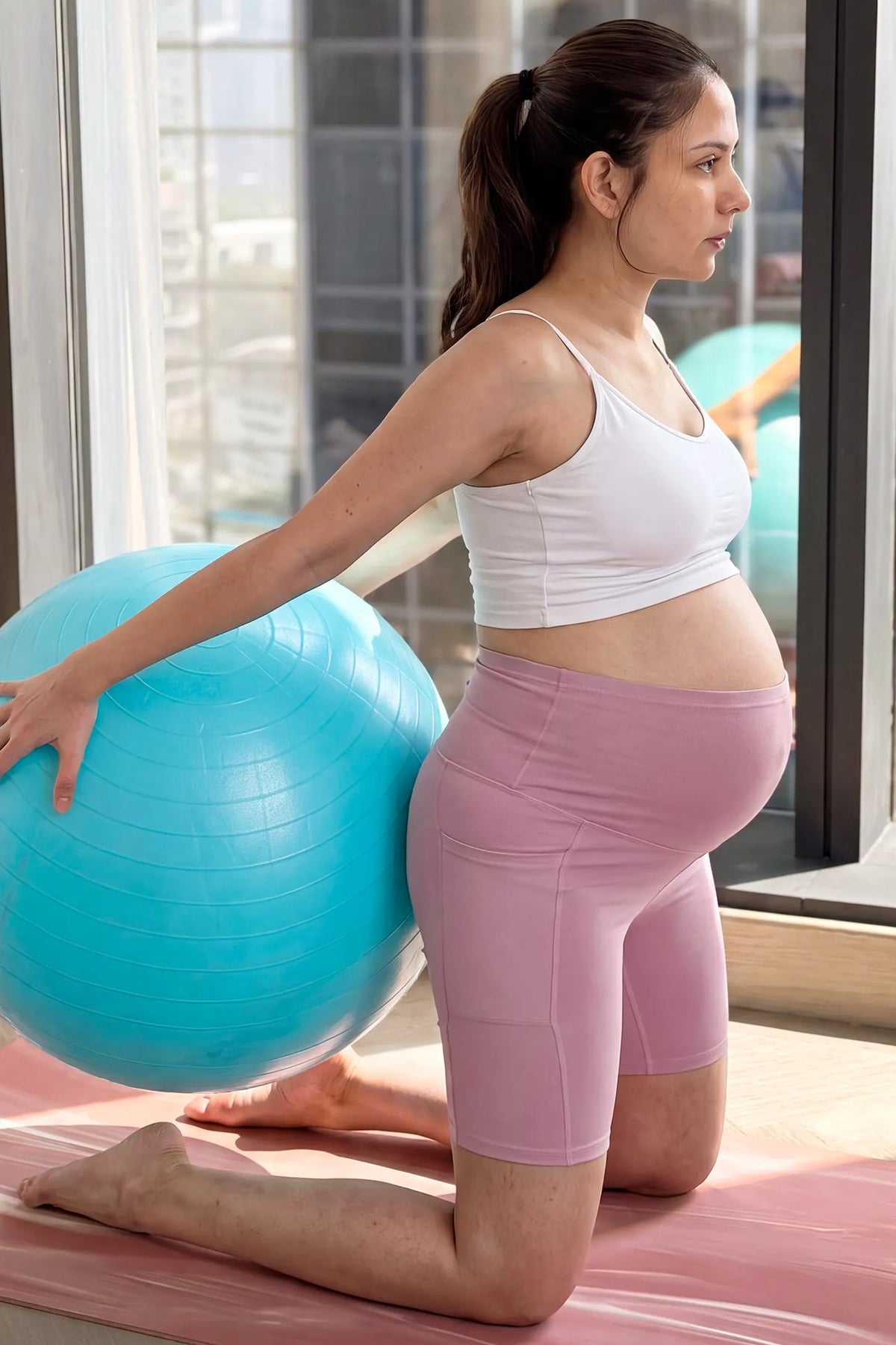 High-Rise Stretch Biker Maternity Shorts High-Rise Stretch Biker Maternity Shorts in Pink featuring comfortable full-belly support, high-stretch jersey feel, and athletic maternity fitness style on a pregnant model with an exercise ball indoors.