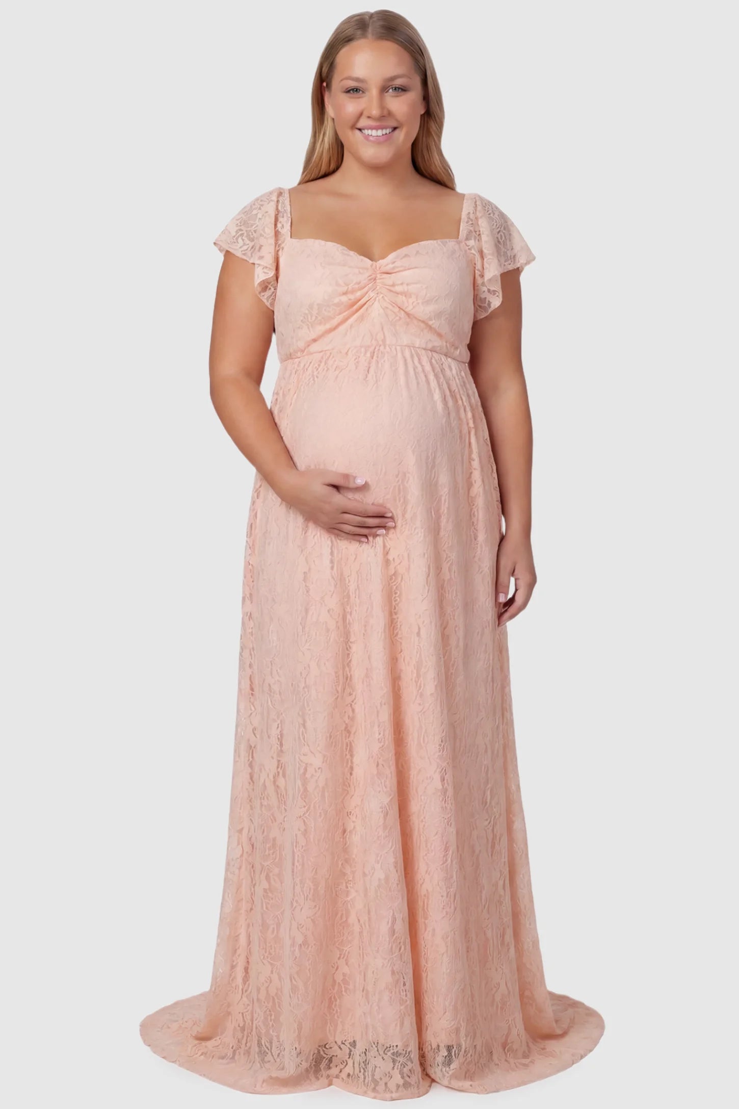 Lace Off Shoulder Maternity Maxi Dress Lace Off Shoulder Maternity Maxi Dress in blush pink featuring a sweetheart neckline with a ruched bust and a full-length skirt, shown on a plus size pregnant model, front view.