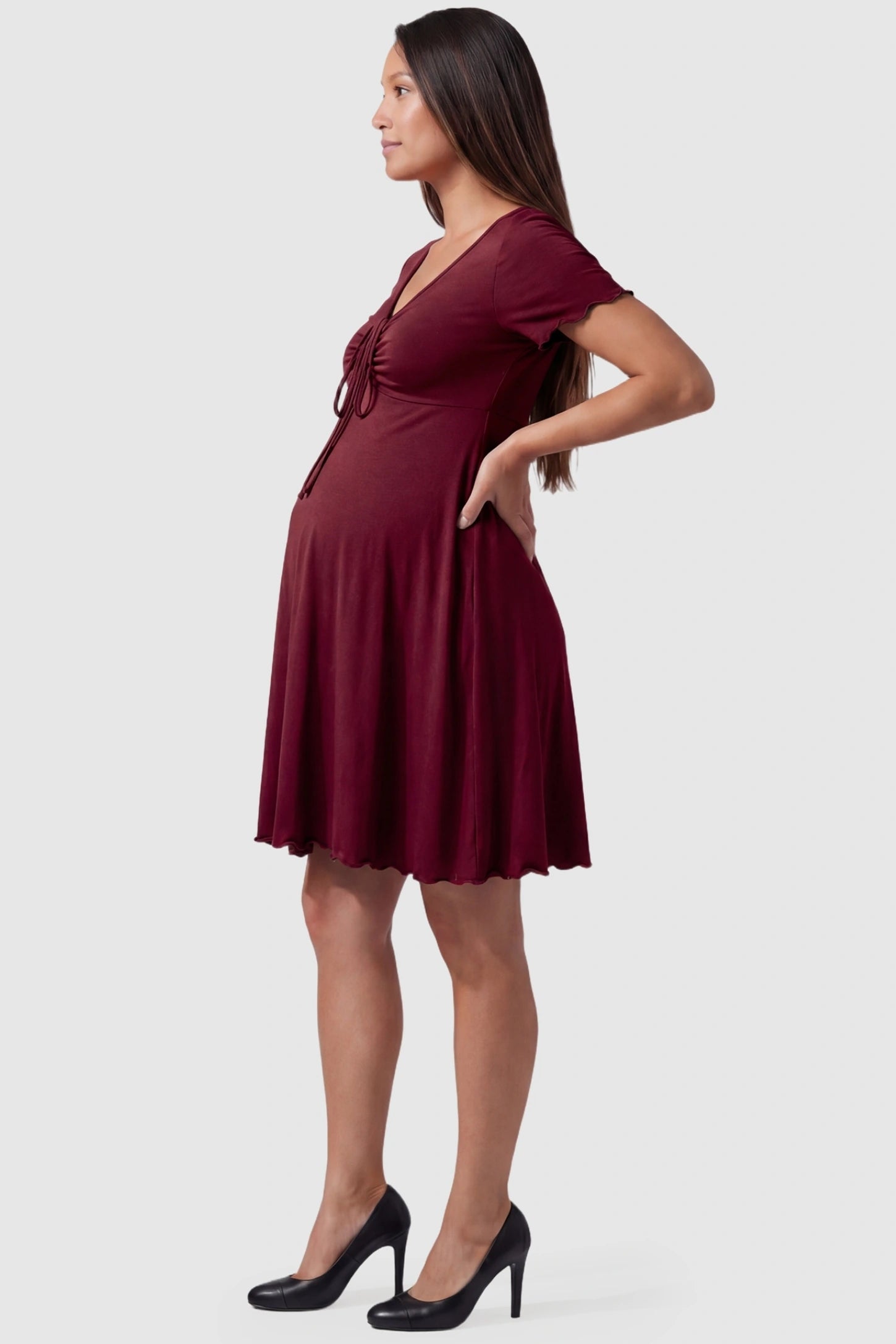Lettuce Trim A-Line Mini Maternity Dress in Burgundy featuring a curve-hugging silhouette on the side profile of a pregnant model.
