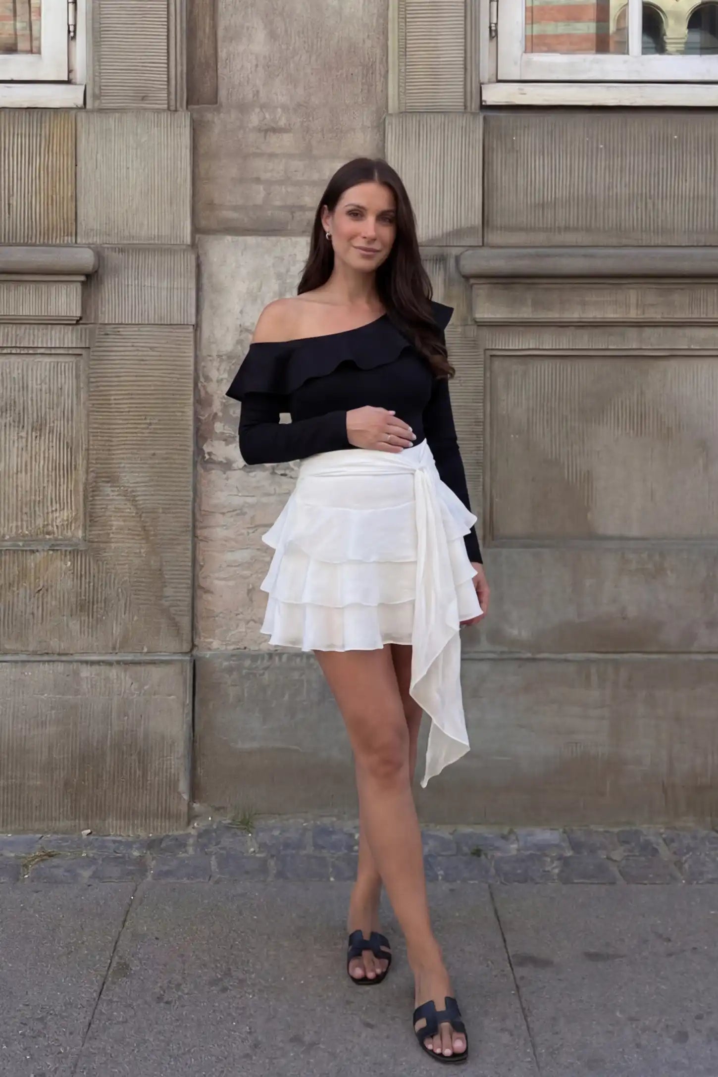 Off-Shoulder Ruffle Trim Maternity Bodysuit Off-shoulder ruffle trim maternity bodysuit in black with long-sleeve and paired with skirt, front view on pregnant model