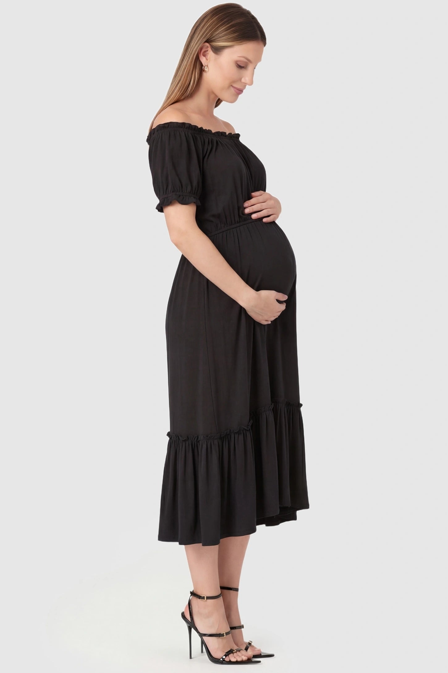 Ruffle Off-The-Shoulder Tiered Maternity Dress Ruffle Off-The-Shoulder Tiered Maternity Dress in Black featuring a tiered ruffle hem on a side-profile pregnant model.