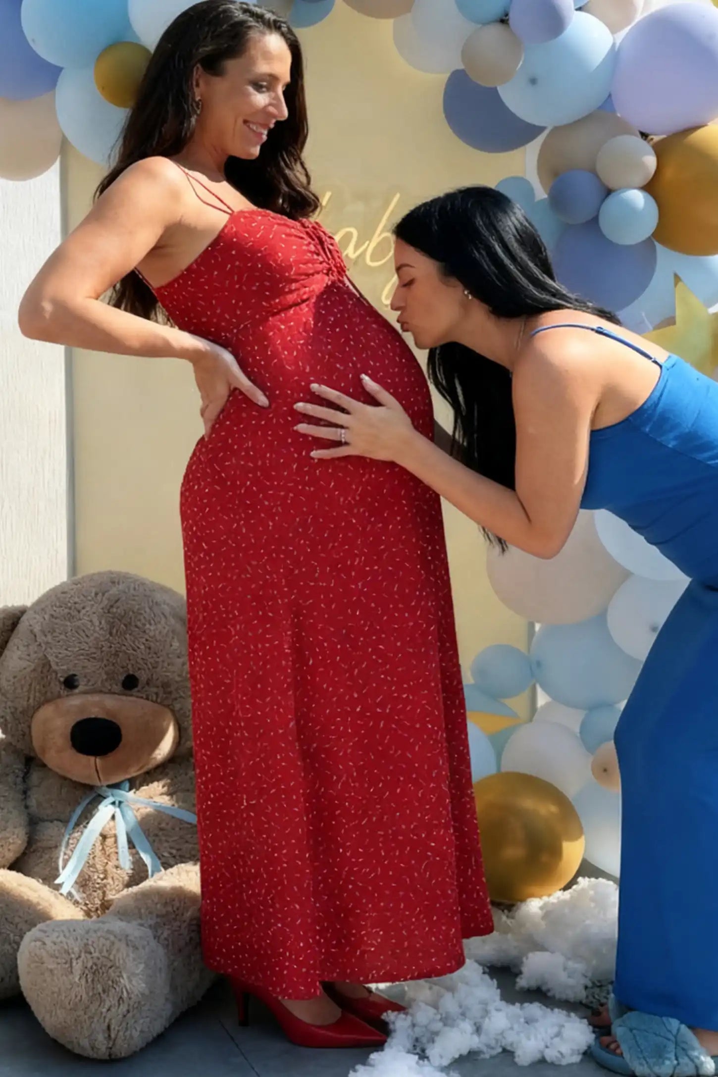 Floral Sweetheart Neck Pleated Maternity Midi Dress Floral Sweetheart Neck Pleated Midi Maternity Dress in Red Ditsy Floral featuring a delicate chiffon drape on a pregnant model while a friend in blue leans in to kiss her baby bump at a baby shower with a balloon backdrop.