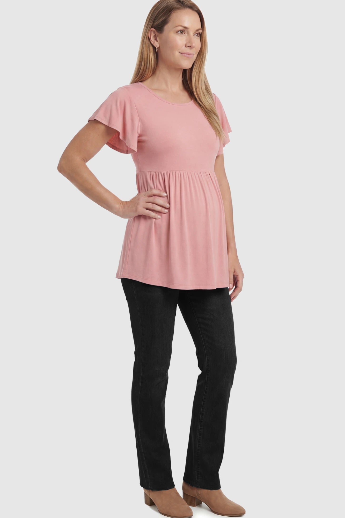 Solid Ruffled-Sleeve Flowy Maternity Top Solid Ruffled-Sleeve Flowy Maternity Top in Pink featuring a pleated empire waist paired with black jeans and boots on a pregnant model in  a full body image.
