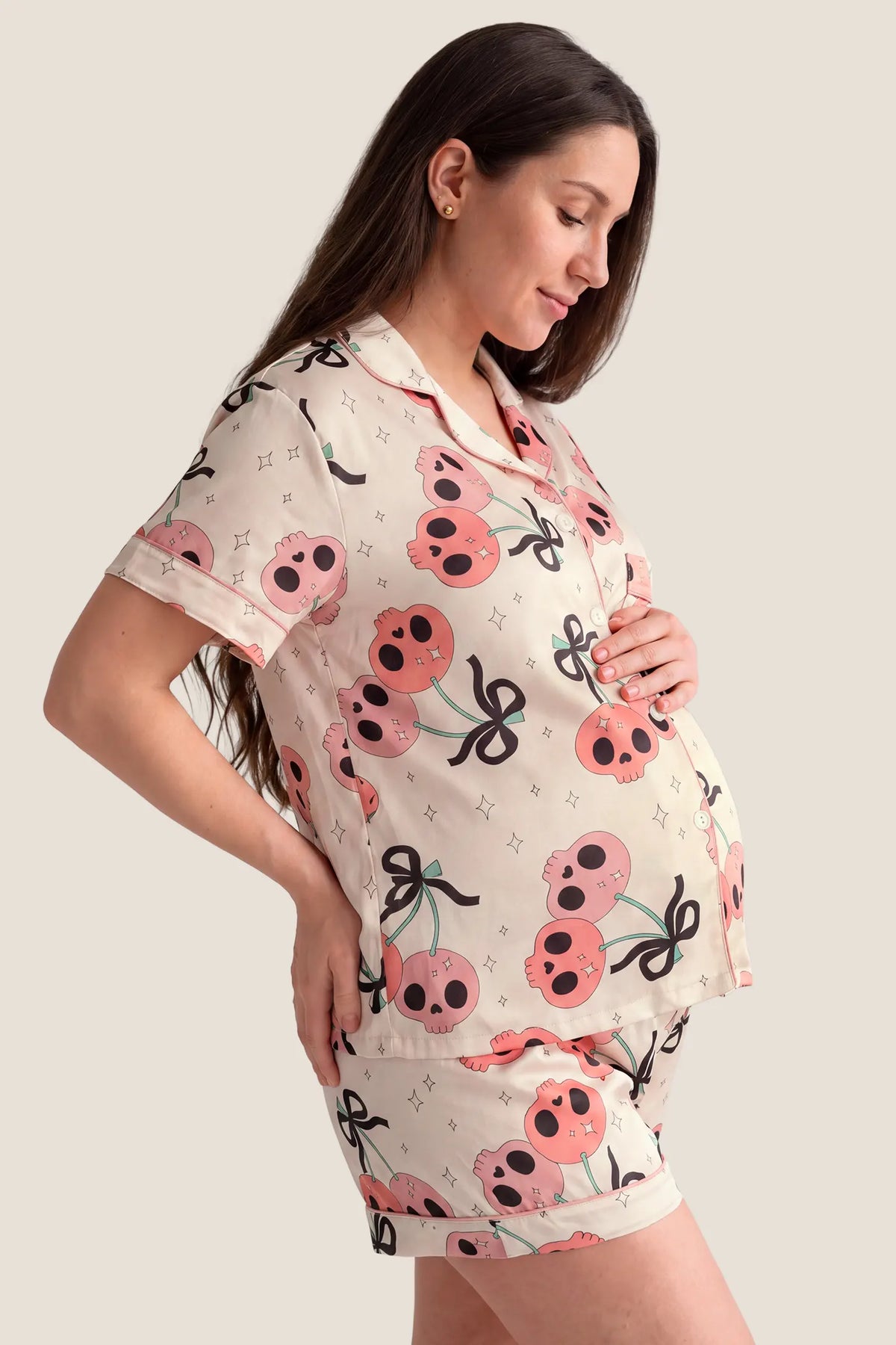 Pink Skull Cherry Button-Up Maternity Pajama Set Pink Skull Cherry Button-Up Maternity Pajama Set in Pink Skull featuring side bump room, short-sleeve collared silhouette, and lightweight Halloween print fabric on a pregnant model, side profile view.