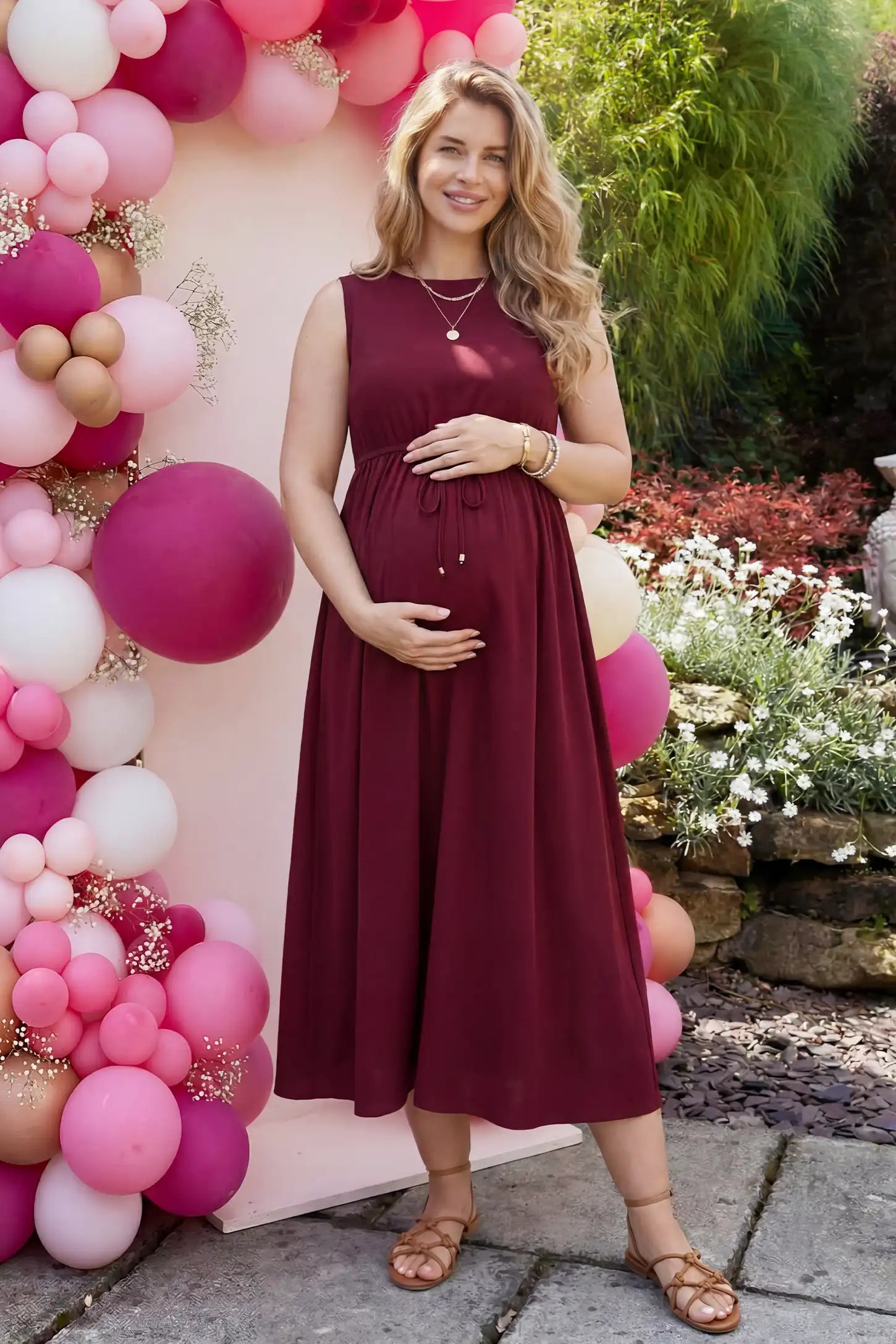 Drawstring Waist Sleeveless Full Skirt Maternity Skater Dress Drawstring Waist Sleeveless Maternity Skater Dress in Burgundy featuring a front profile bump view on a pregnant model outdoors.