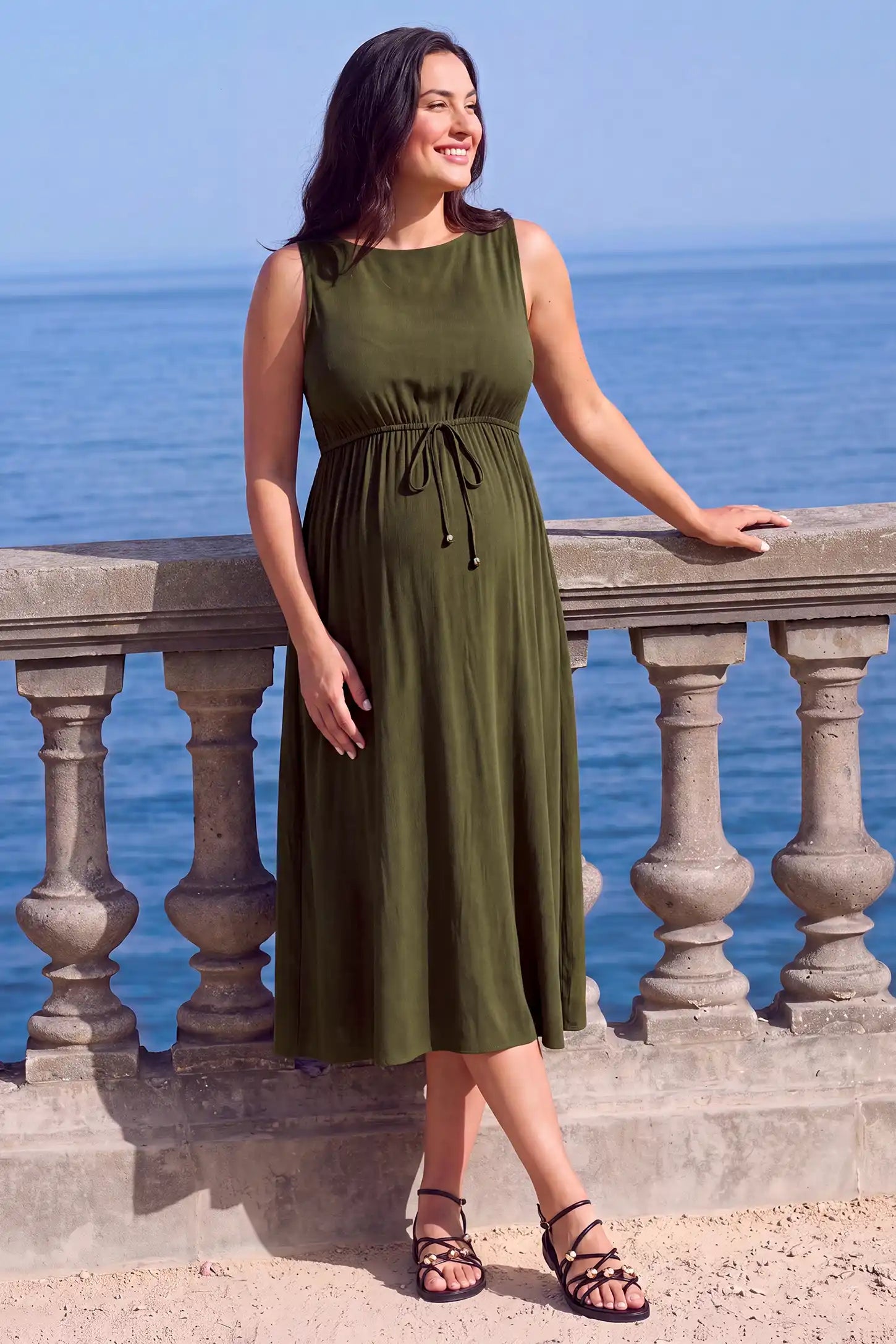 Drawstring Waist Sleeveless Full Skirt Maternity Skater Dress Drawstring Waist Sleeveless Maternity Skater Dress in green featuring a side profile bump view on a pregnant model outdoors.