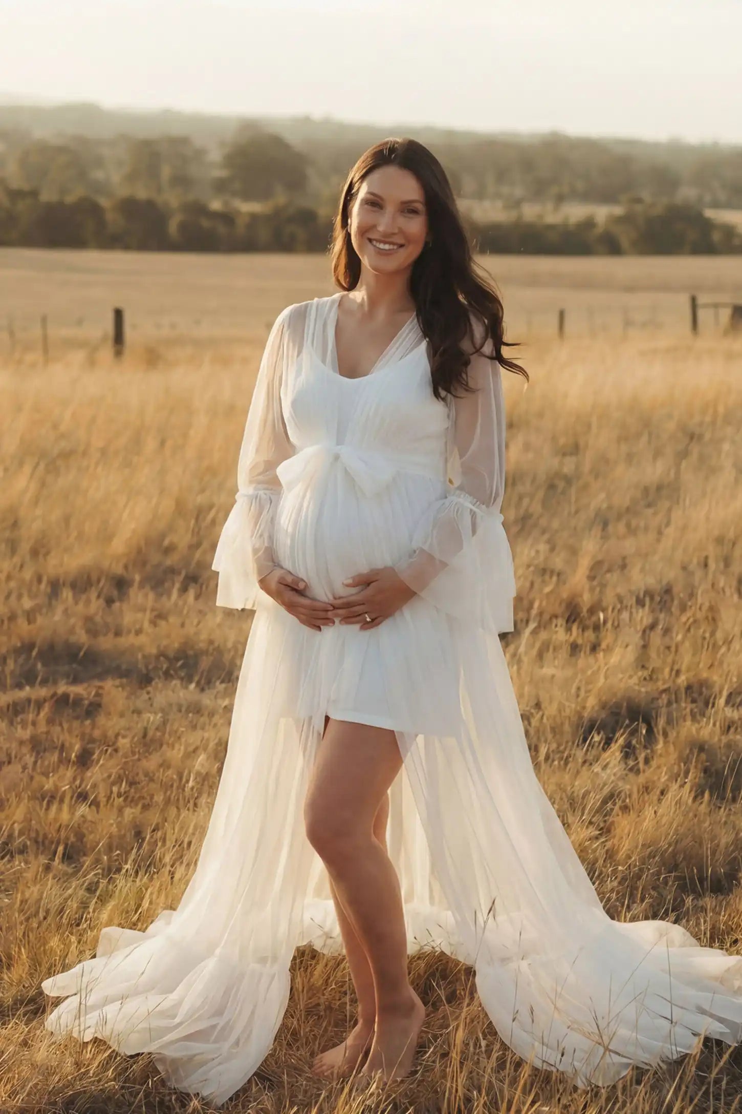 Sheer Tulle Deep V-Neck Tiered Maternity Maxi Gown Sheer tulle deep V-neck tiered maternity maxi gown in white with deep V-neckline and statement bow waist, editorial lifestyle view