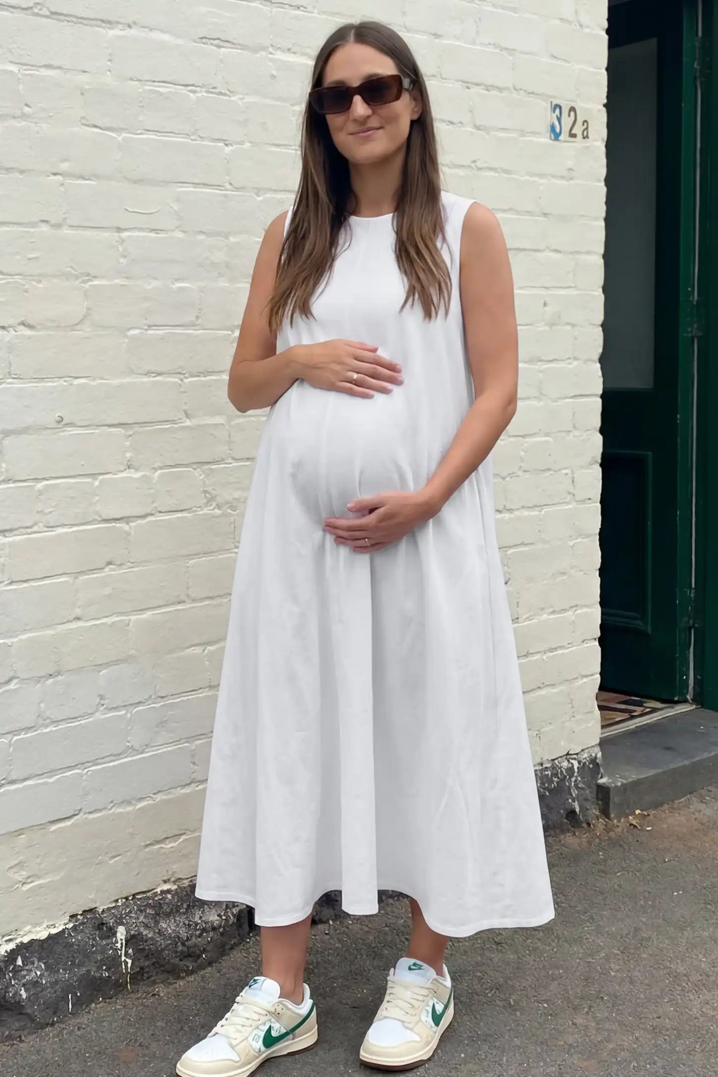 Sleeveless Pocketed Longline Hem Maternity Midi Dress Sleeveless pocketed longline hem maternity midi dress in white with sleeveless design and longline hem, front view on pregnant model