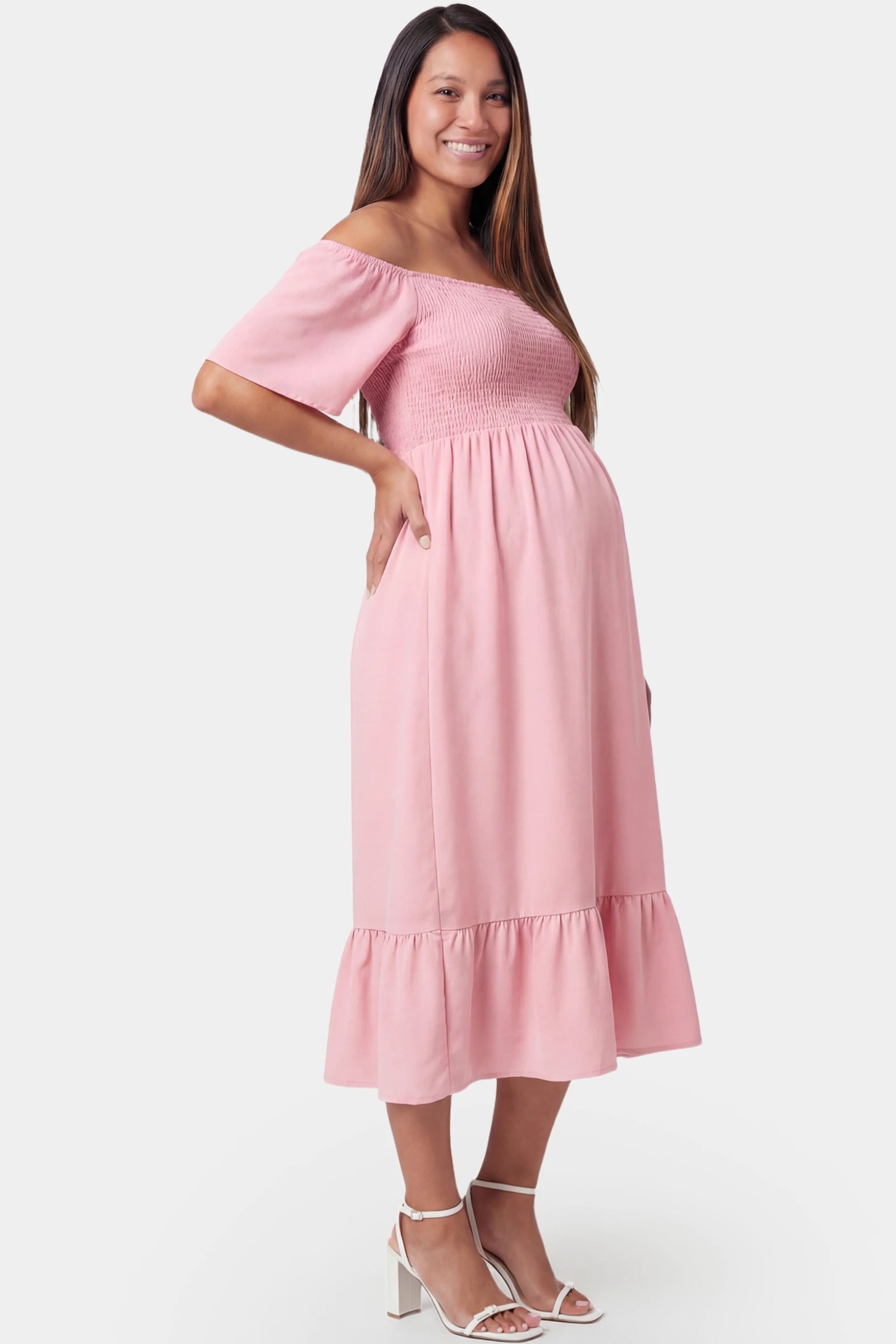 Off-shoulder smocked short sleeve A-line midi maternity dress in pink featuring delicate ruffle detail in side view on a pregnant model