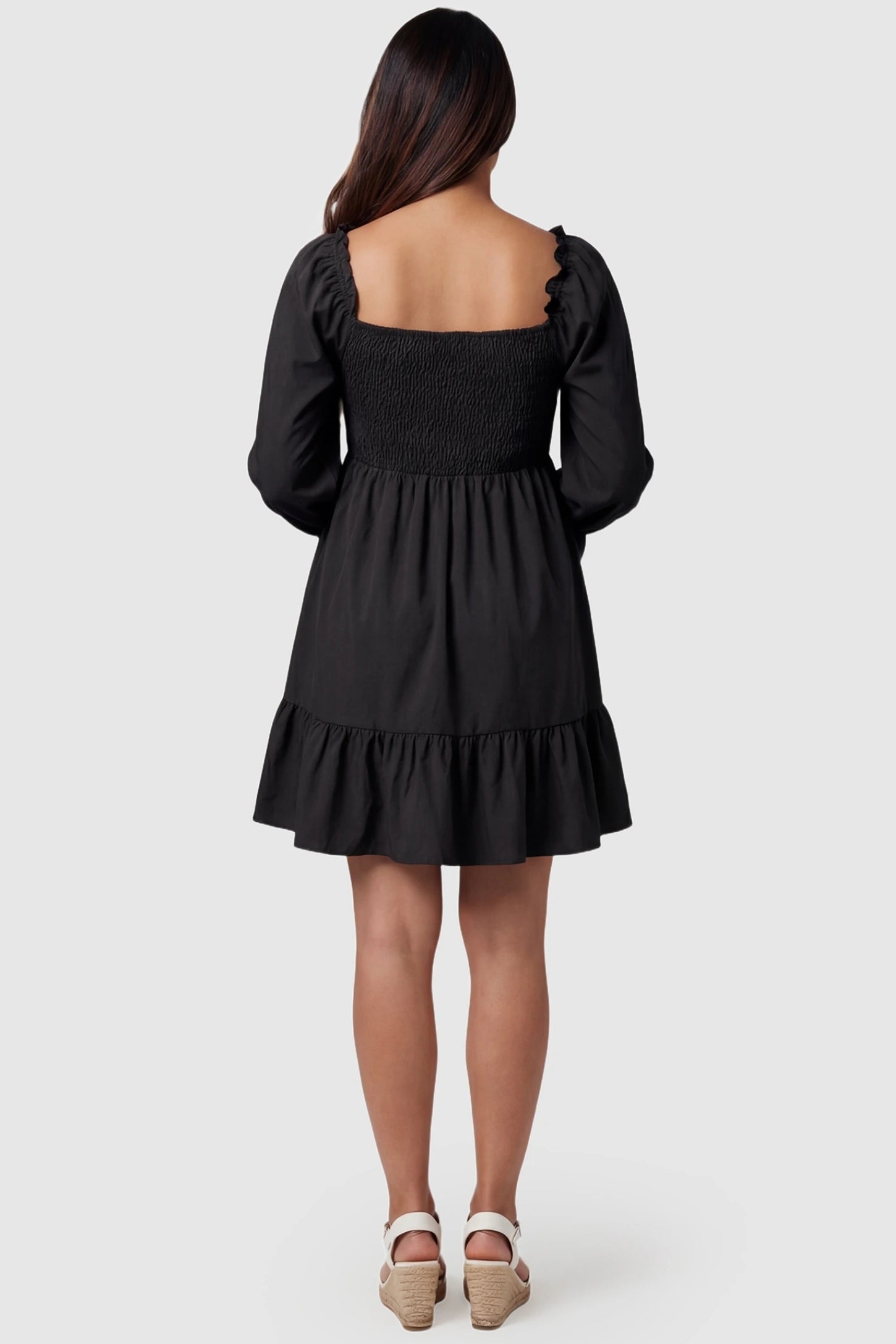 Smocked Chiffon A-Line Maternity Mini Dress Smocked Chiffon A-Line Maternity Mini Dress in Black showing the ruffled hem and flared sleeves on a pregnant model, back view.