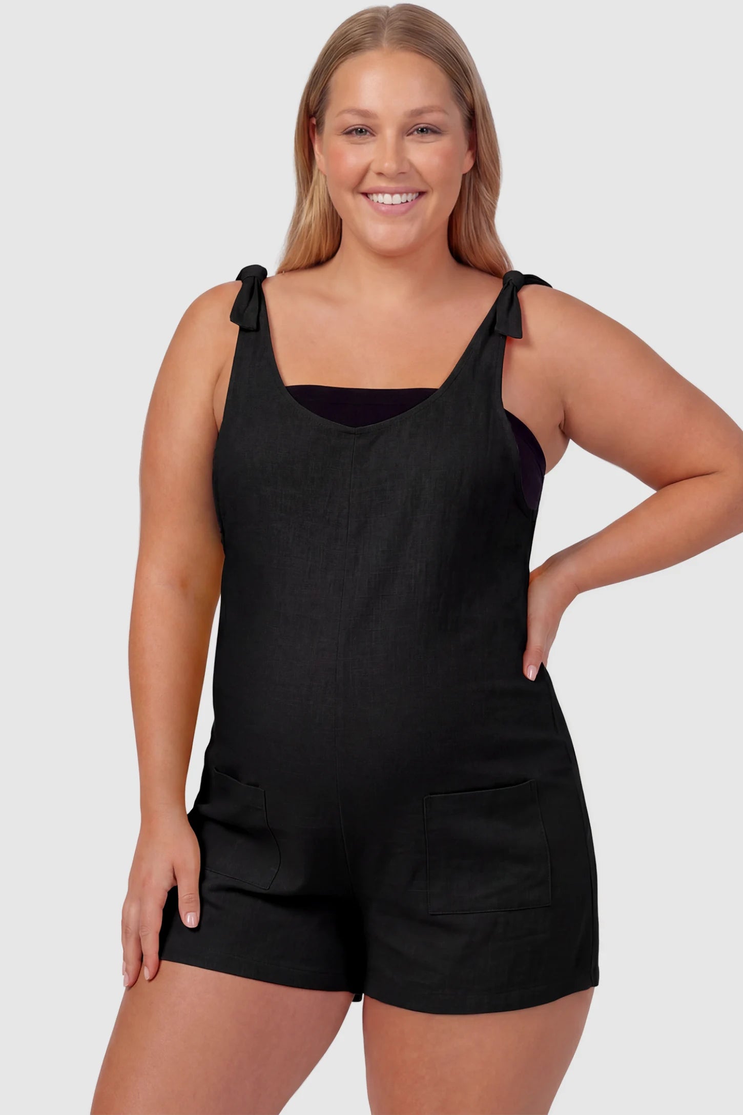 Tie-Strap Sleeveless Pocketed Maternity Romper Tie-strap sleeveless pocketed maternity romper in black with adjustable tie straps and scoop neckline, front view on plus-size model