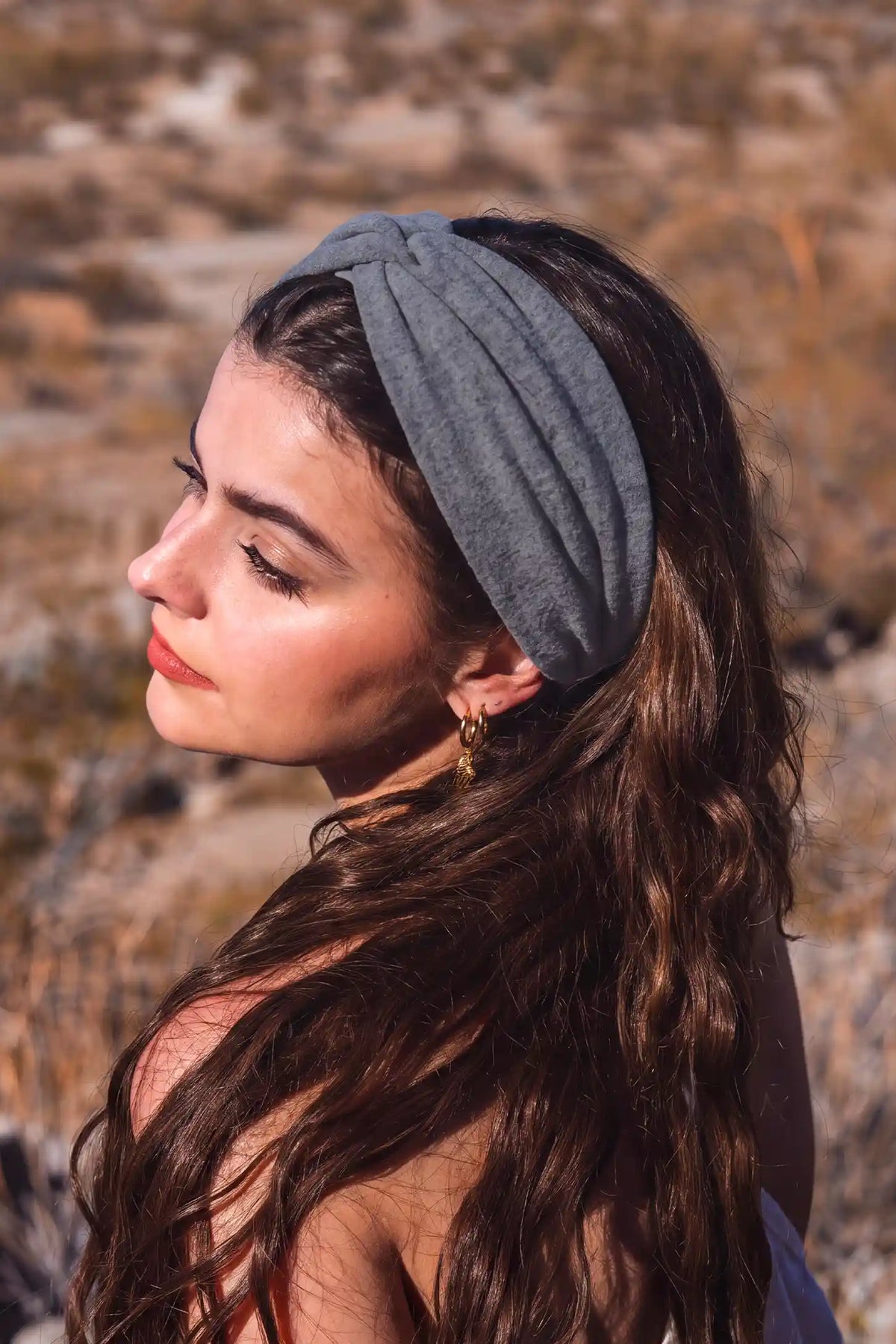 Wide Crossed Headband Wide Crossed Headband in Heather Grey featuring a soft stretch ribbed fabric and polished crossed twist detail on a brunette model outdoors, side profile view.