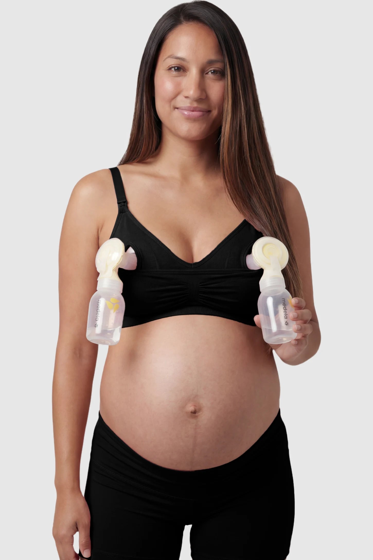 Seamless Ruched Front Maternity Nursing Bra Seamless Ruched Front Maternity Nursing Bra in black with hands-free pumping bottles attached, showing the innovative crossover design on a smiling pregnant model showing front.