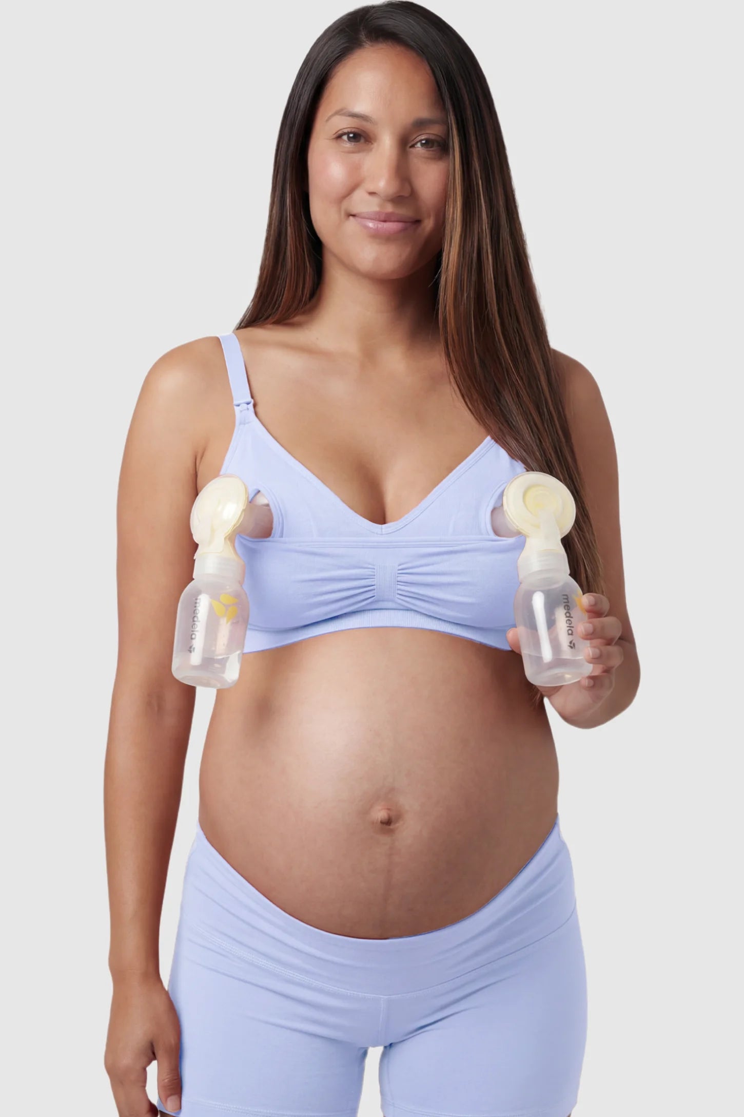 Seamless Ruched Front Maternity Nursing Bra Seamless Ruched Front Maternity Nursing Bra in blue with hands-free pumping bottles attached, showing the innovative crossover design on a smiling pregnant model showing front.