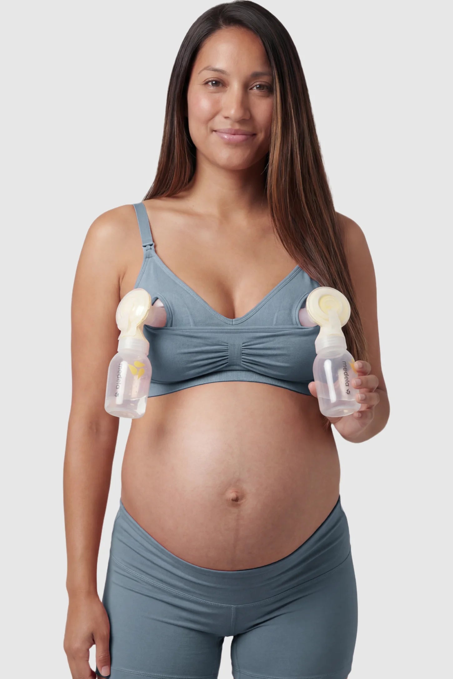 Seamless Ruched Front Maternity Nursing Bra Seamless Ruched Front Maternity Nursing Bra in gray with hands-free pumping bottles attached, showing the innovative crossover design on a smiling pregnant model showing front.