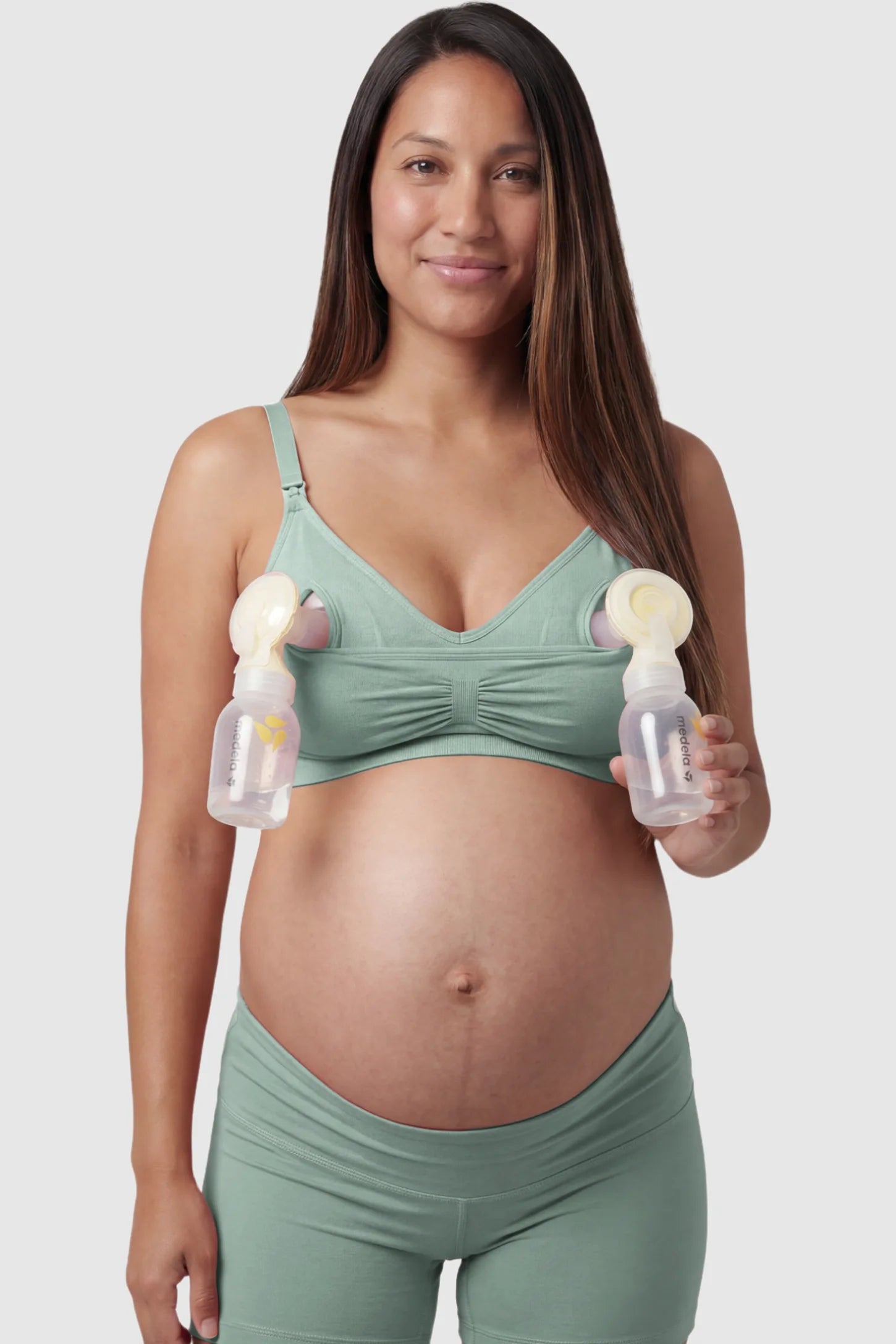 Seamless Ruched Front Maternity Nursing Bra Seamless Ruched Front Maternity Nursing Bra in green with hands-free pumping bottles attached, showing the innovative crossover design on a smiling pregnant model showing front.