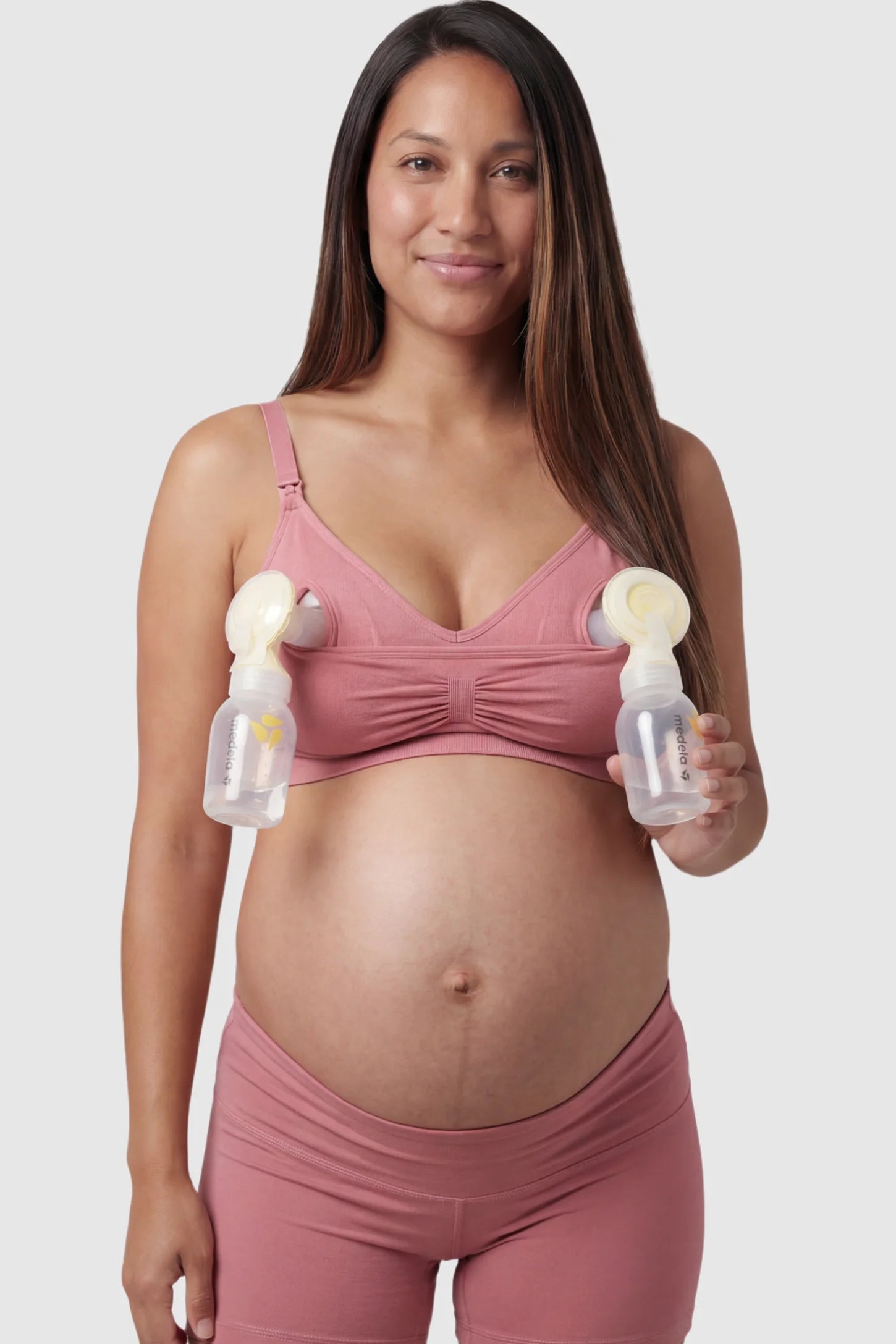 Seamless Ruched Front Maternity Nursing Bra Seamless Ruched Front Maternity Nursing Bra in pink with hands-free pumping bottles attached, showing the innovative crossover design on a smiling pregnant model showing front.