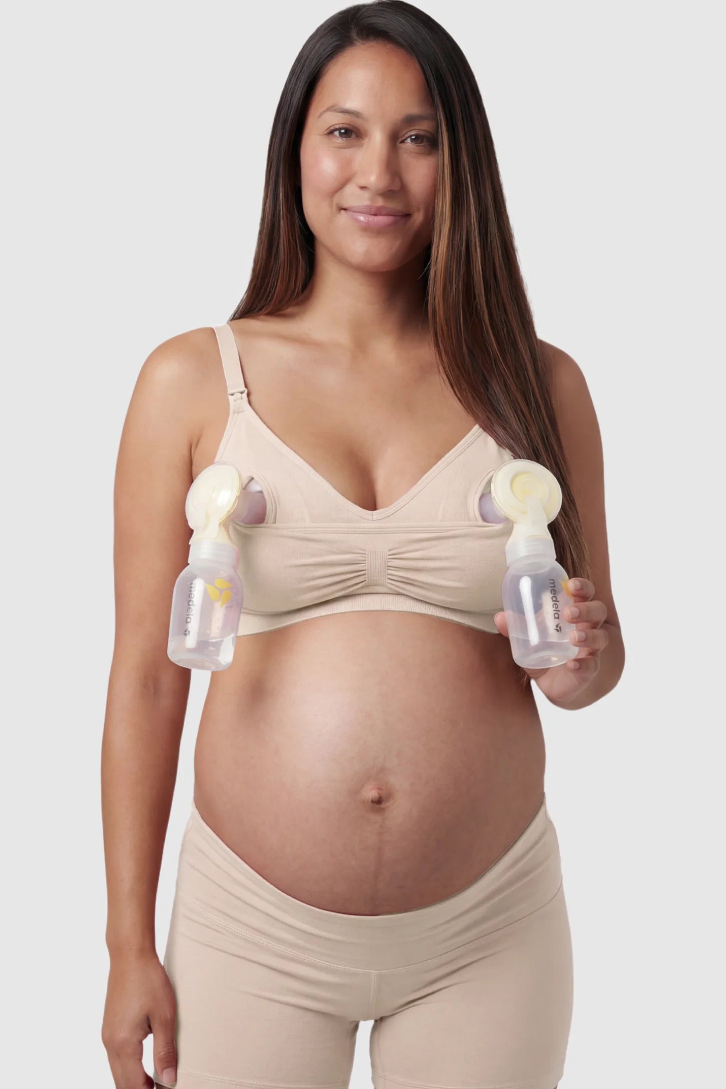 Seamless Ruched Front Maternity Nursing Bra Seamless Ruched Front Maternity Nursing Bra in skin with hands-free pumping bottles attached, showing the innovative crossover design on a smiling pregnant model showing front.