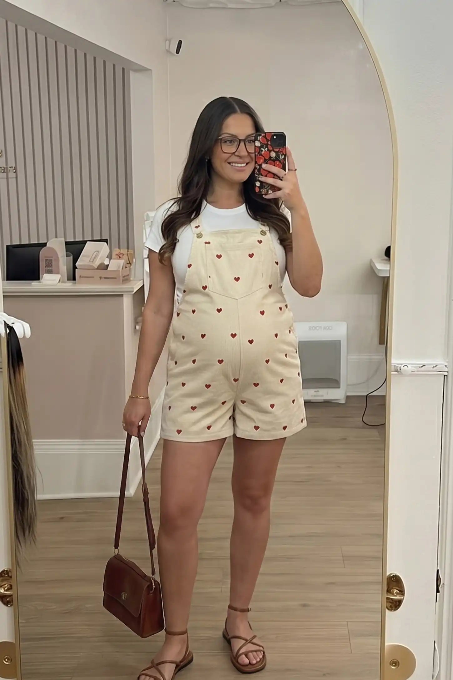 Adjustable Waist Front Pocket Maternity Short Overalls Pregnant woman taking mirror selfie wearing cream adjustable waist front pocket maternity short overalls with embroidered hearts and gold buttons, paired with white t-shirt, brown leather crossbody bag and strappy sandals
