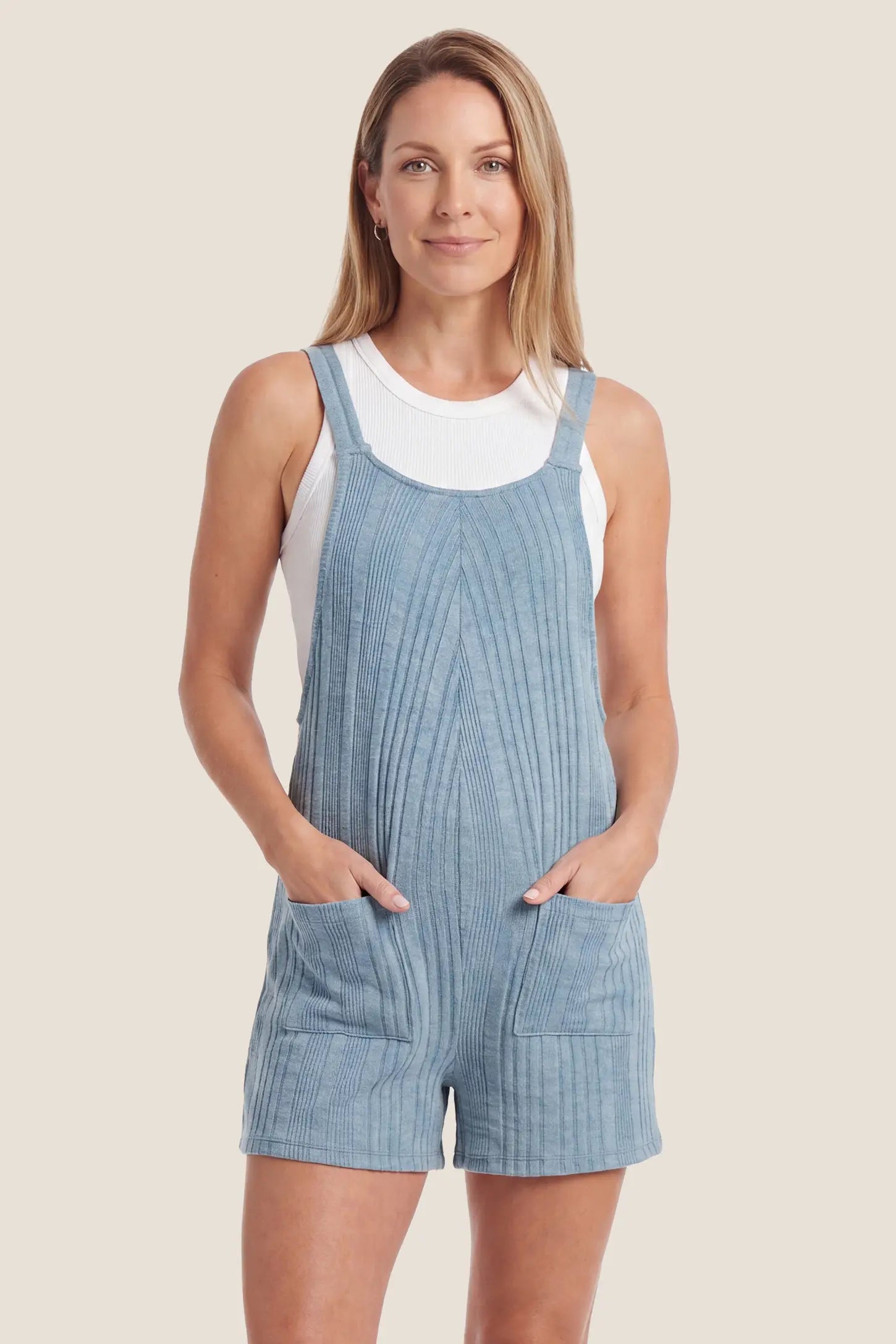 Ribbed Pocket Knit Maternity Romper Woman wearing blue ribbed square-neck short overalls romper with two front patch pockets, hands in pockets, paired with white tank top, on beige background

