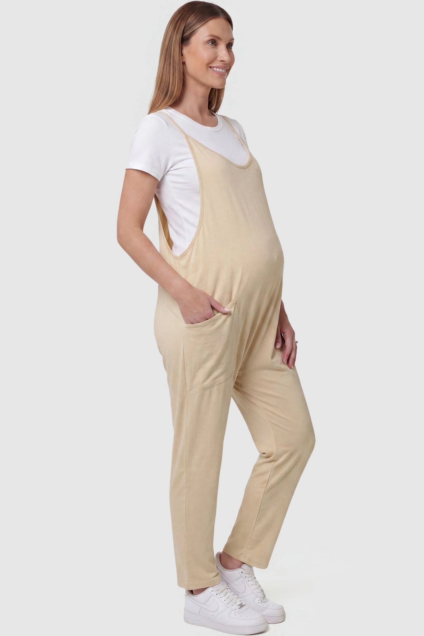Adjustable Strap Pocketed Relaxed Maternity Jumpsuit Adjustable Strap Pocketed Maternity Jumpsuit in Beige with functional side pockets on a pregnant model, side view.