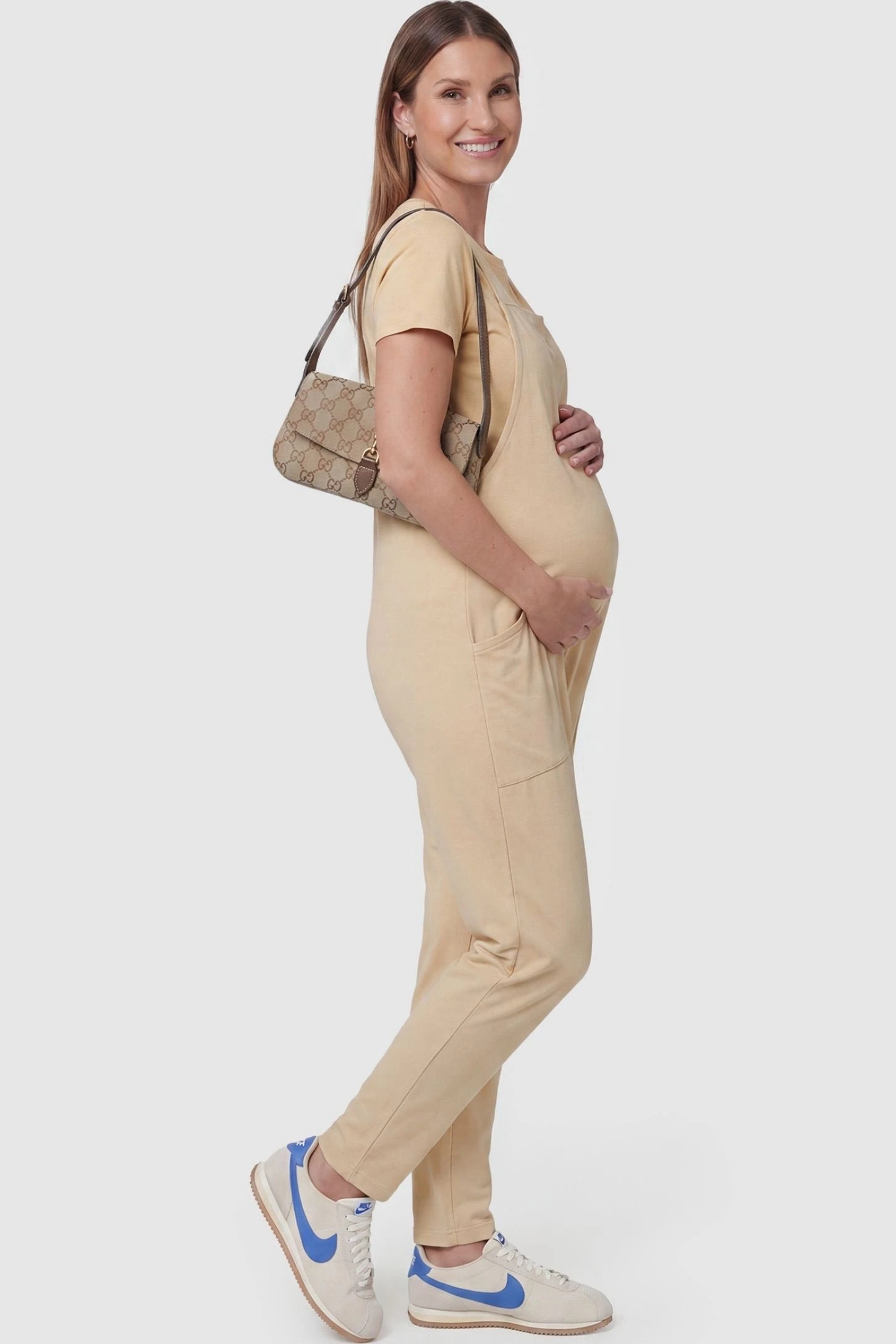 Adjustable Strap Pocketed Relaxed Maternity Jumpsuit Adjustable Strap Pocketed Maternity Jumpsuit in Beige with functional side pockets on a pregnant model, side view.