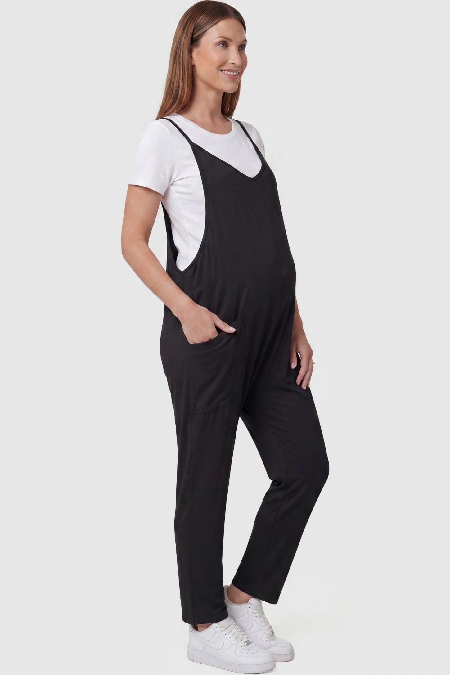 Adjustable Strap Pocketed Relaxed Maternity Jumpsuit Adjustable Strap Pocketed Maternity Jumpsuit in Black with functional side pockets on a pregnant model, side view.