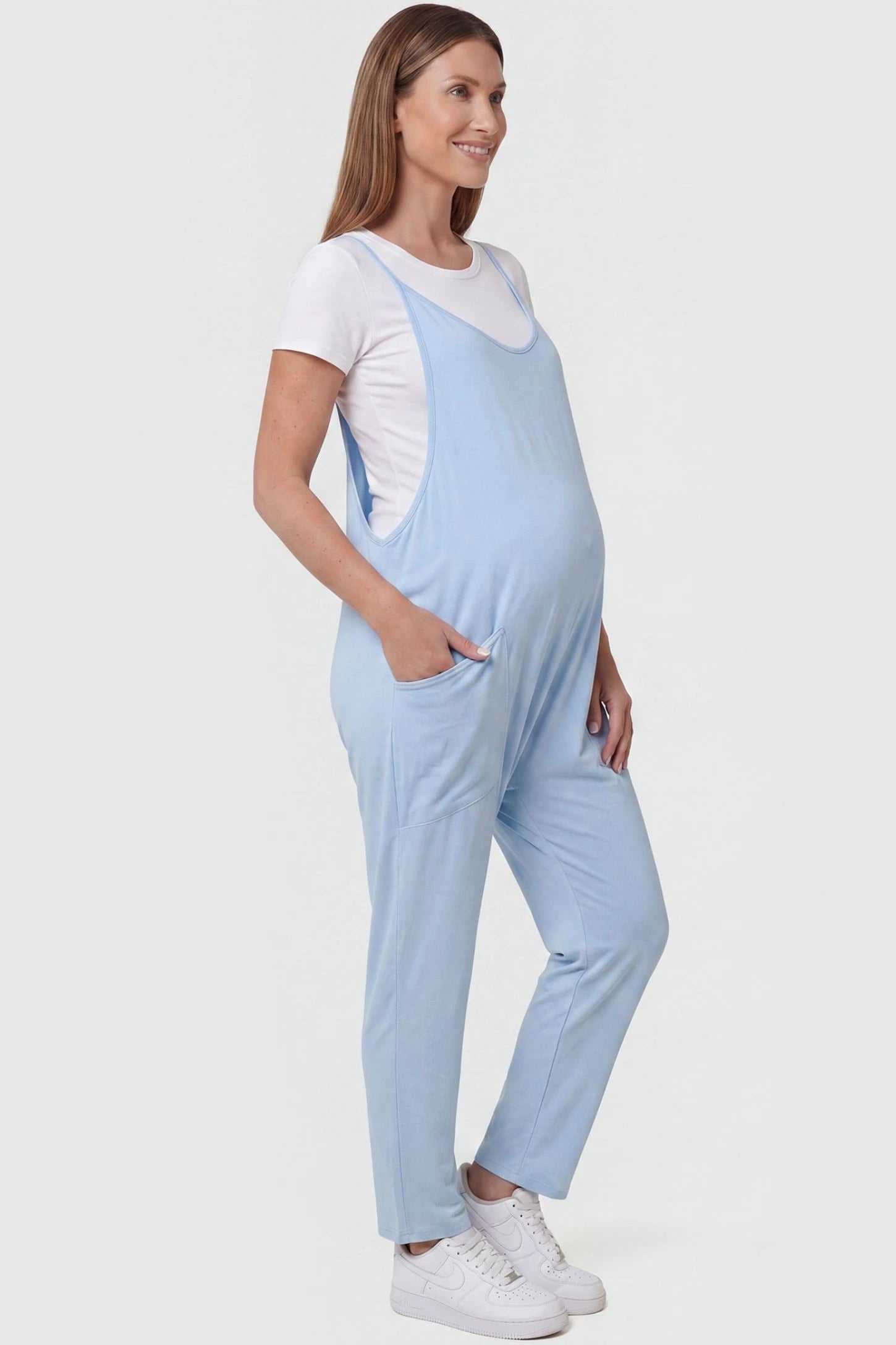 Adjustable Strap Pocketed Relaxed Maternity Jumpsuit Adjustable Strap Pocketed Maternity Jumpsuit in Light Blue with functional side pockets on a pregnant model, side view.