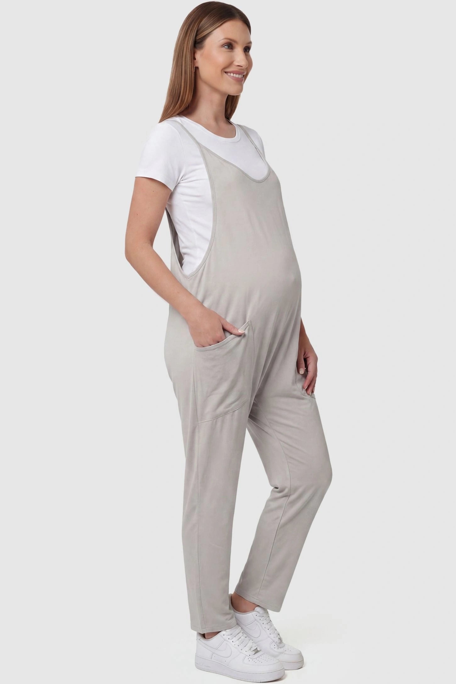 Adjustable Strap Pocketed Relaxed Maternity Jumpsuit Adjustable Strap Pocketed Maternity Jumpsuit in Light Gray with functional side pockets on a pregnant model, side view.