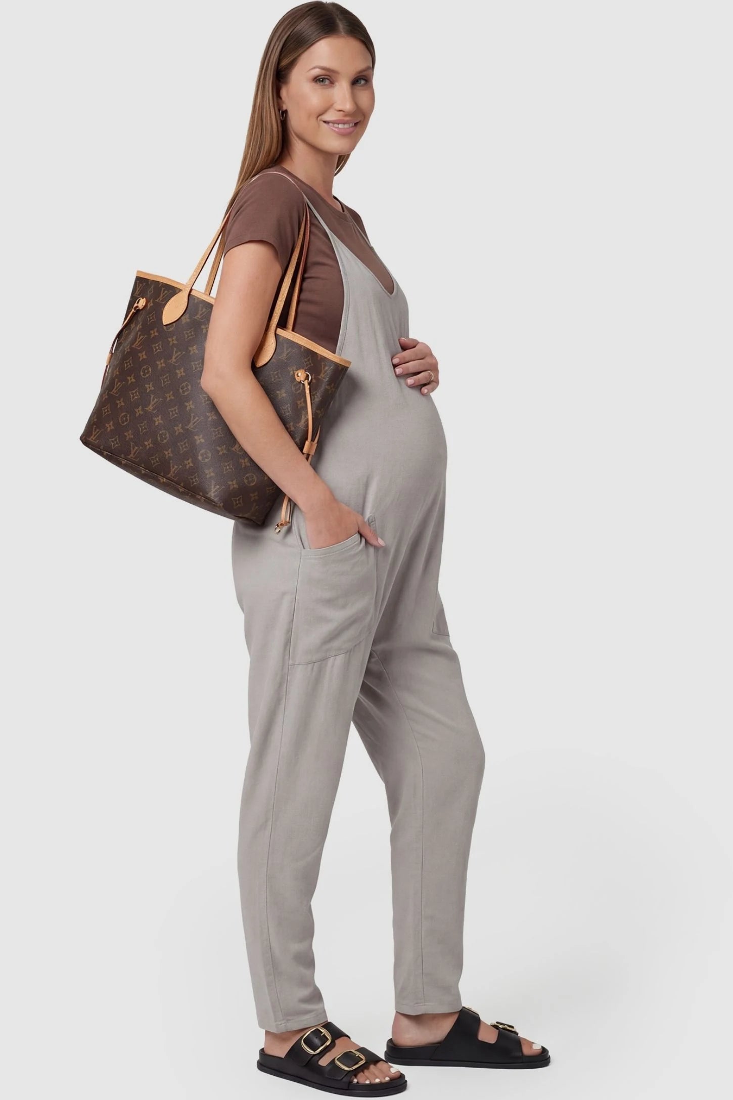 Adjustable Strap Pocketed Relaxed Maternity Jumpsuit Adjustable Strap Pocketed Maternity Jumpsuit in Light Gray with functional side pockets on a pregnant model, side view.