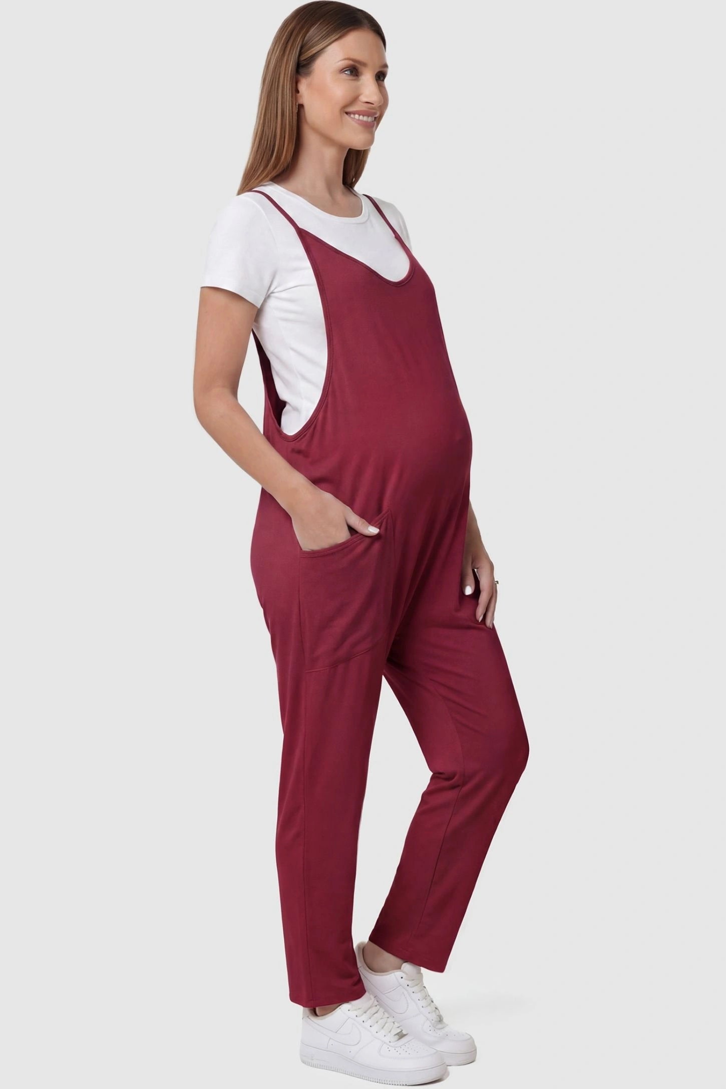 Adjustable Strap Pocketed Relaxed Maternity Jumpsuit Adjustable Strap Pocketed Maternity Jumpsuit in Wine Red with functional side pockets on a pregnant model, side view.