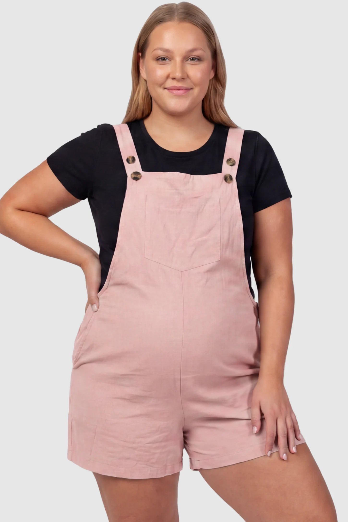 Adjustable Waist Pocketed Maternity Short Overalls Adjustable Waist Pocketed Maternity Short Overalls in Pink featuring a bib pocket on a plus size pregnant model, front view.
